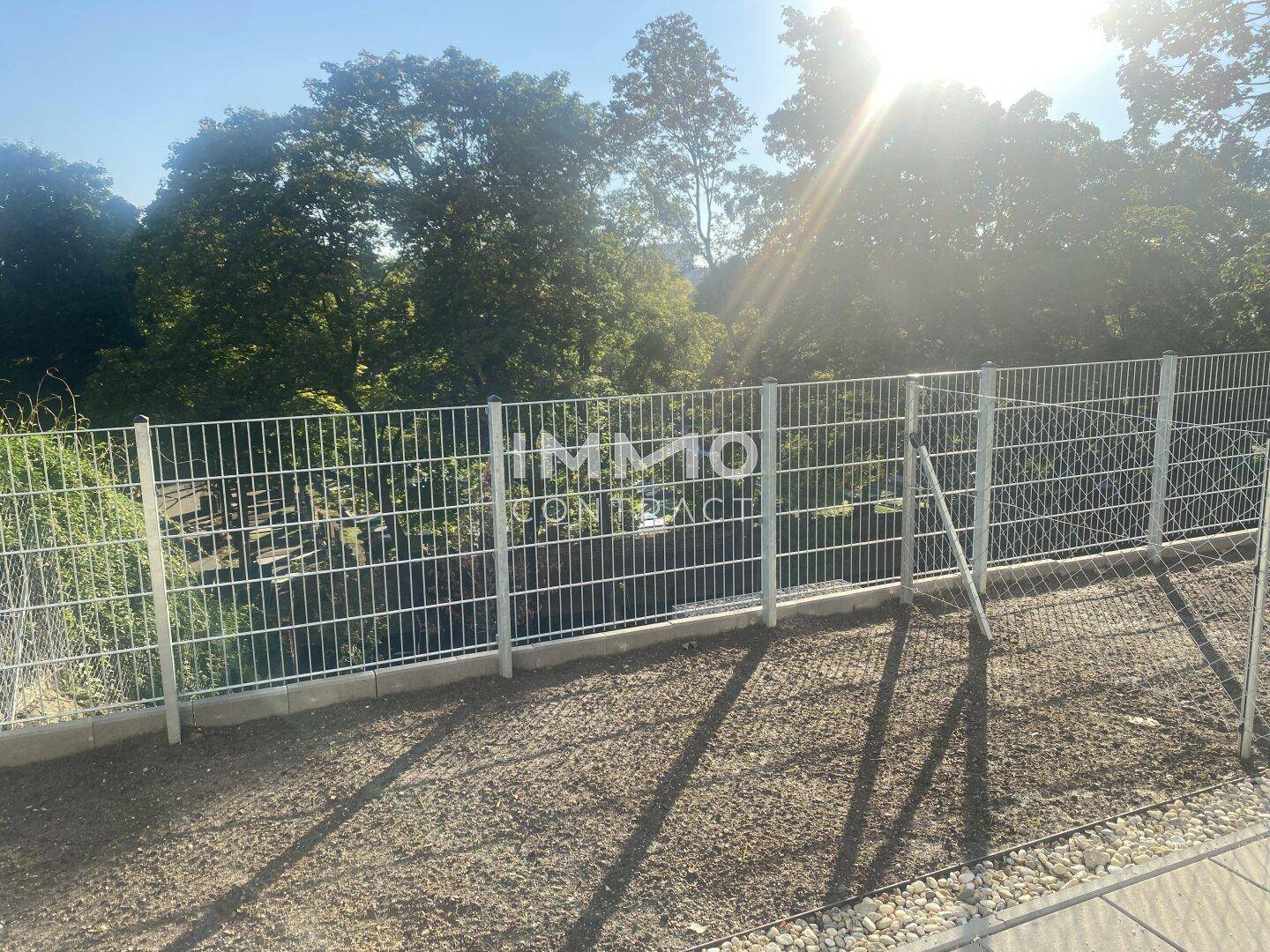 Eingezäunter Außenbereich mit Kies und Blick auf Bäume unter strahlendem Sonnenschein.