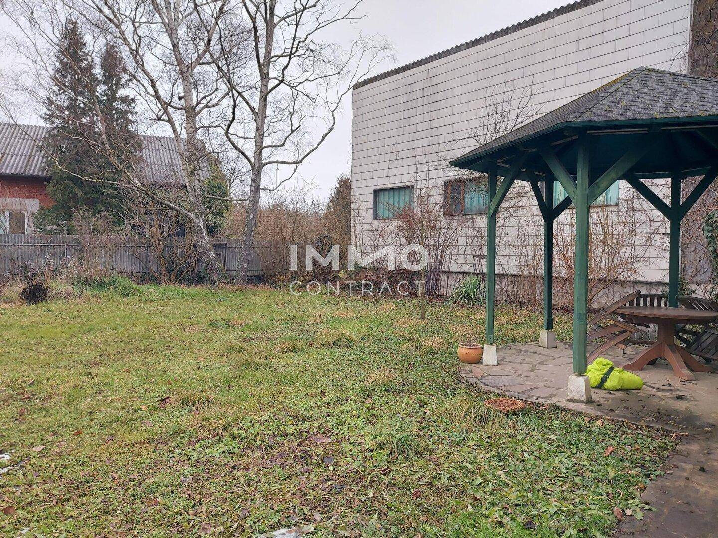 Gartenbereich mit Rasenfläche, Laube und kahlen Bäumen im Hintergrund.