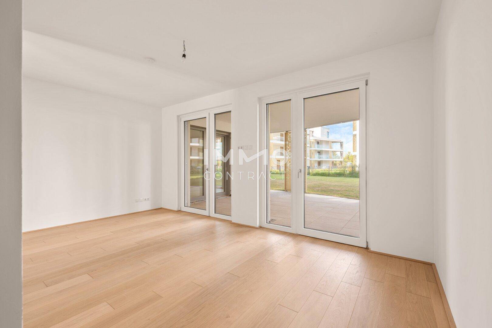 Geräumiger Wohnraum mit hellem Parkettboden und großen Fenstern, die viel Licht hereinlassen.