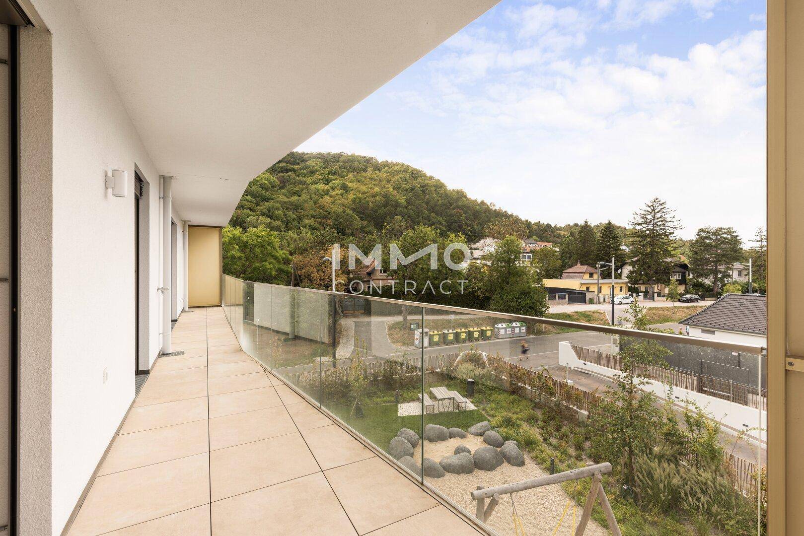 Langer Balkon mit Glasgeländer und Blick auf die grüne Umgebung und einen Spielplatz.