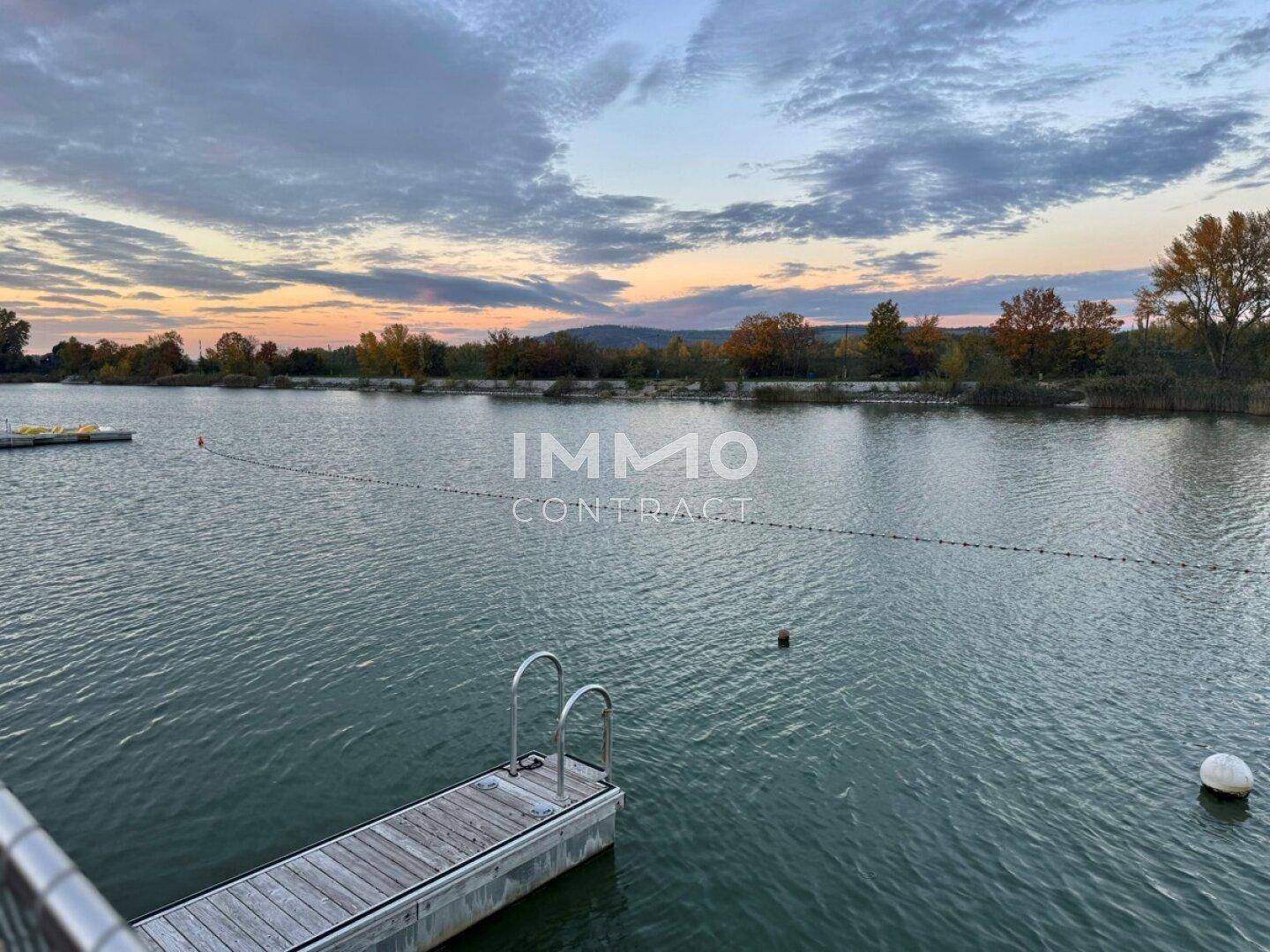Ruhiger See bei Sonnenuntergang mit herbstlicher Baumkulisse und weitem Himmel.