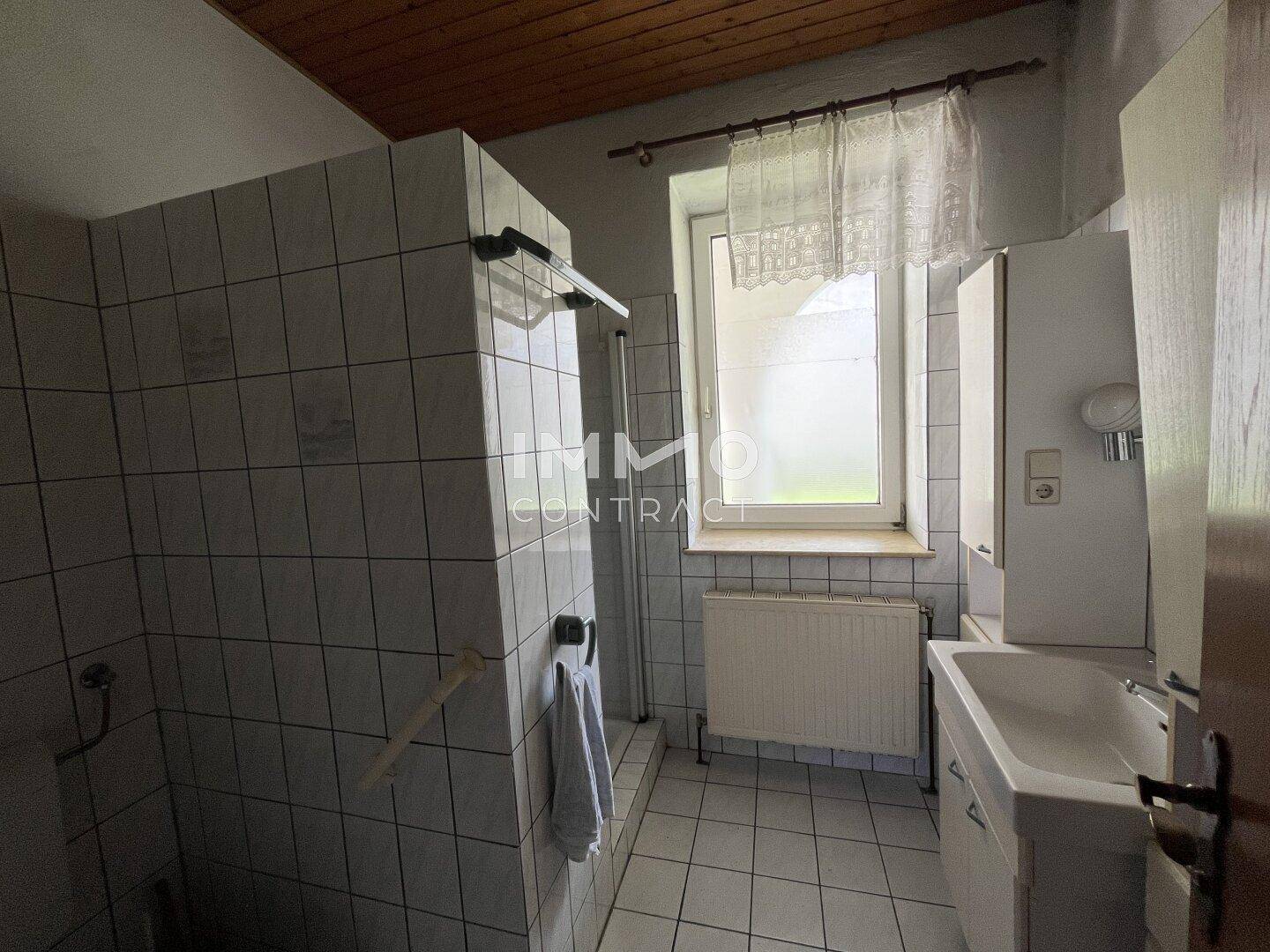 Badezimmer mit Duschbereich, Waschbecken und Fenster, ausgestattet mit weißen Fliesen.