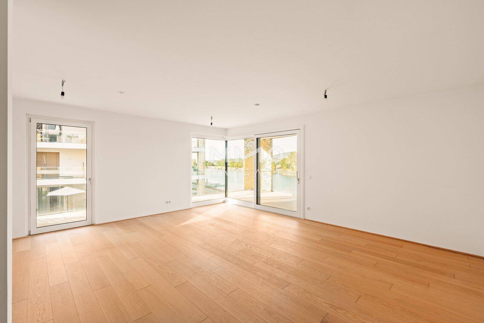Helles, leeres Wohnzimmer mit neuem Holzparkettboden und großen Fenstern mit Seeblick.