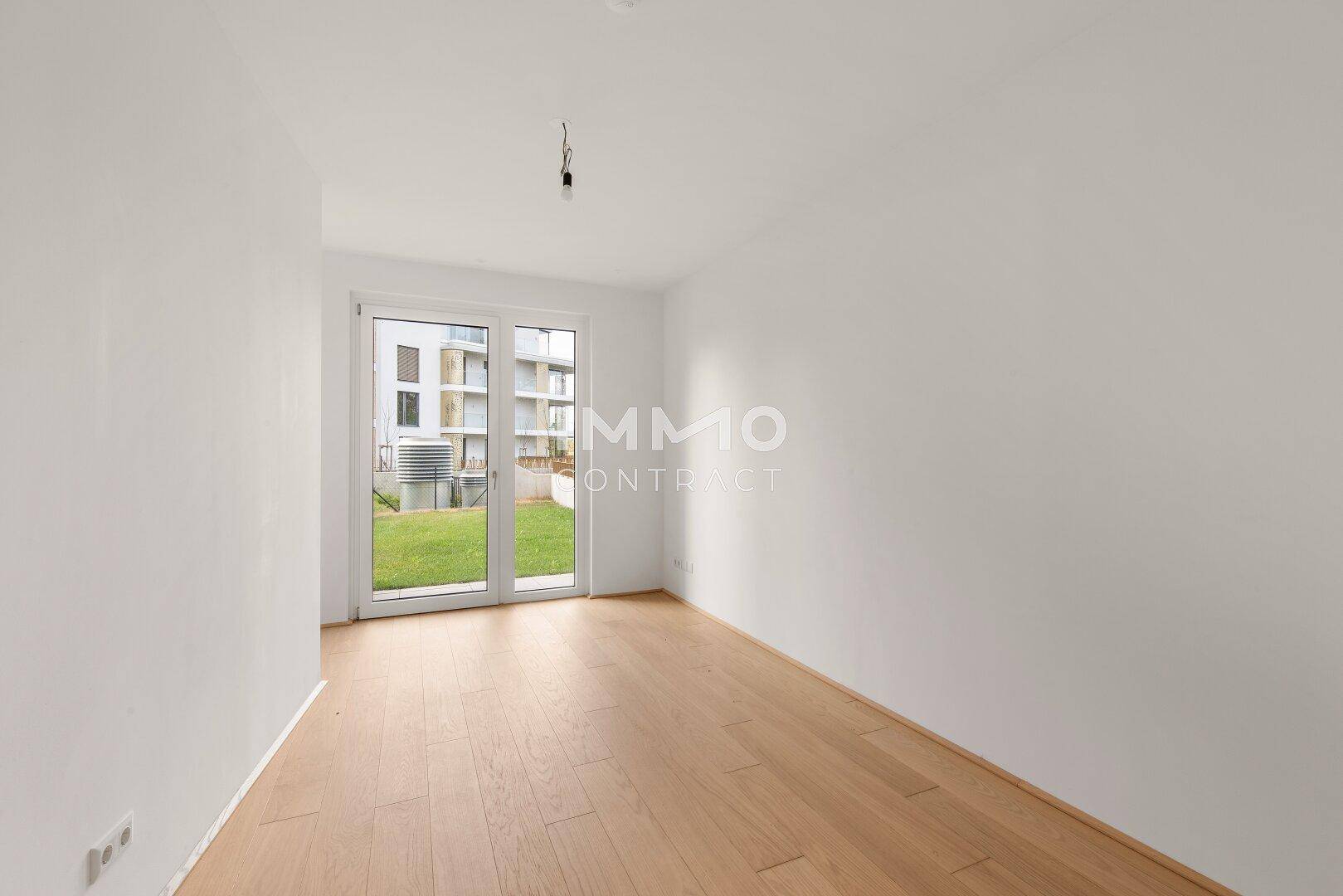 Heller, leerer Raum mit hellem Holzparkett und Zugang zu einer kleinen Rasenfläche im Freien.
