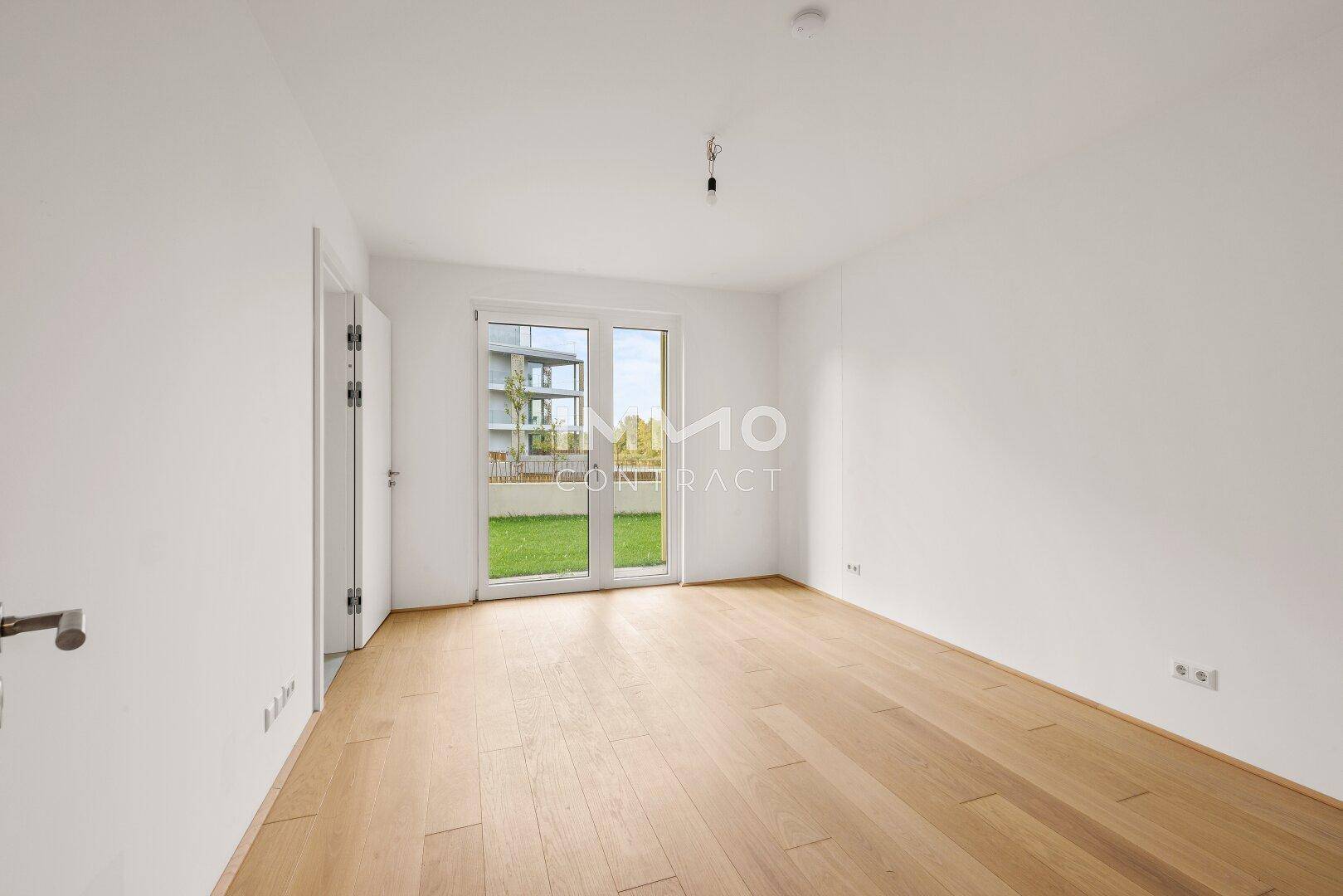 Heller, leerer Raum mit hellem Holzparkett und direktem Zugang zu einer kleinen Grünfläche.