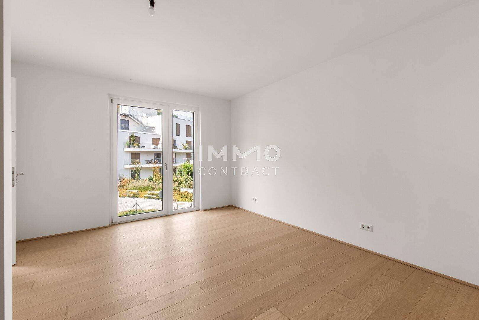 Helles Schlafzimmer mit Parkettboden und großem Fenster, das viel Tageslicht hereinlässt.