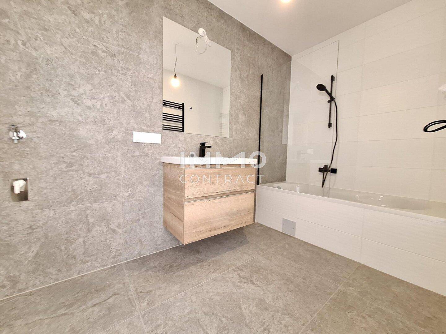 Modernes Badezimmer mit grauen Fliesen, schwebendem Waschtisch, Badewanne und beheiztem Handtuchhalter.