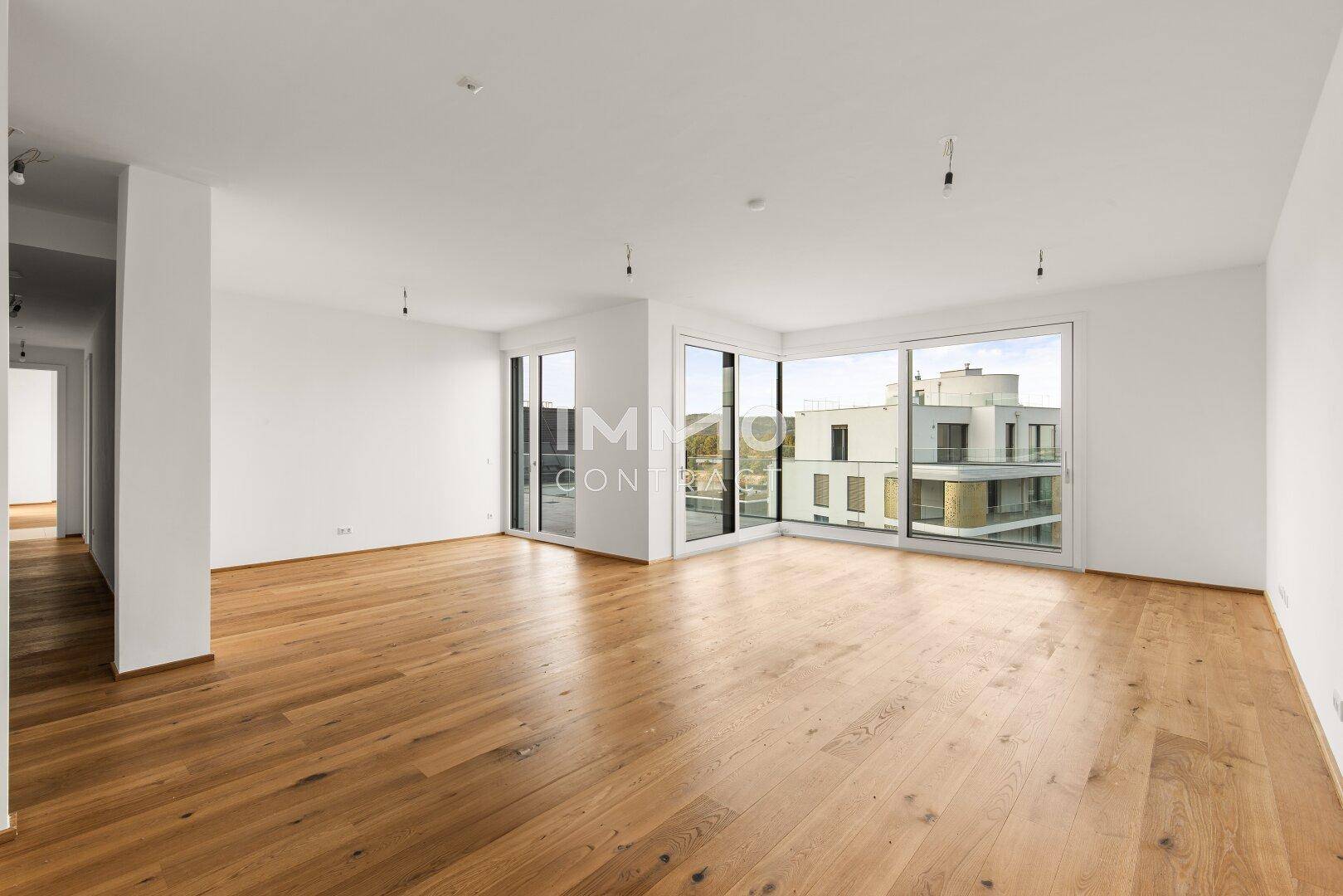 Geräumiges, leeres Wohnzimmer mit Holzboden und großen Fenstern, die viel Licht hereinlassen.