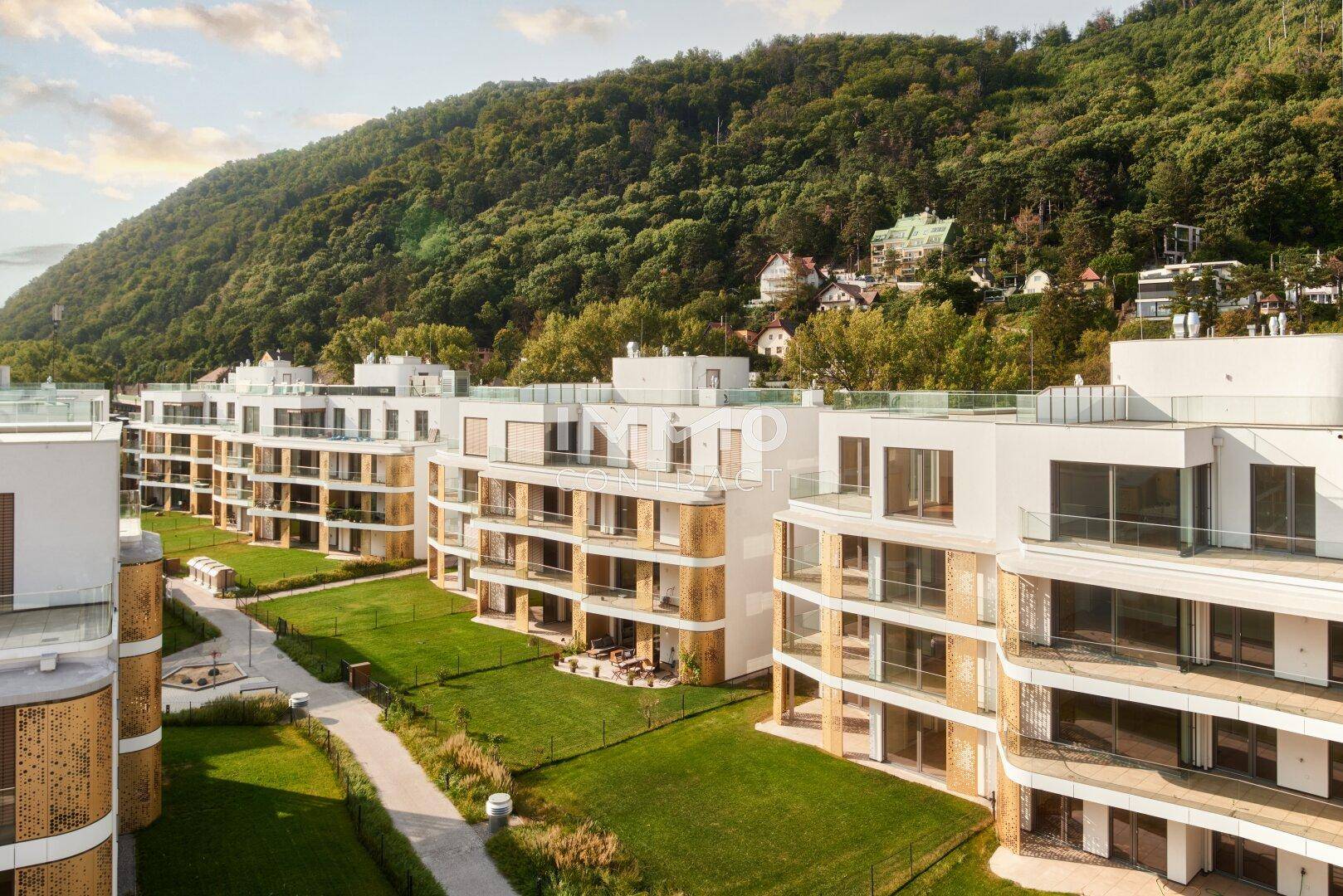 Neubauwohnungen mit modernen Fassaden, Balkonen und gepflegten Grünflächen vor bewaldetem Hang.