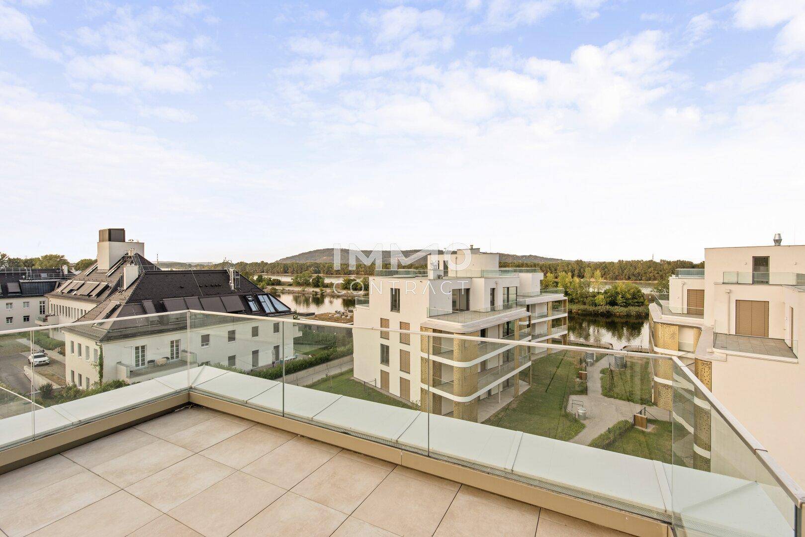 Große Dachterrasse mit Fliesenboden und Glasgeländer, mit Blick auf die Umgebung und das Wasser.