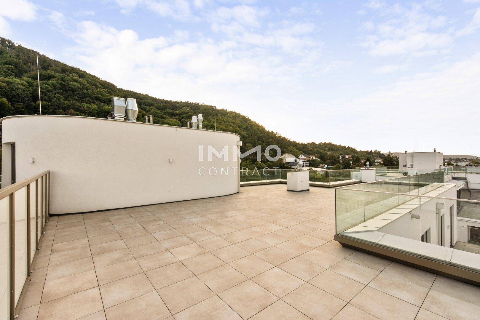 Große Dachterrasse mit Fliesenboden und moderner Glasbrüstung, umgeben von grünen Hügeln.