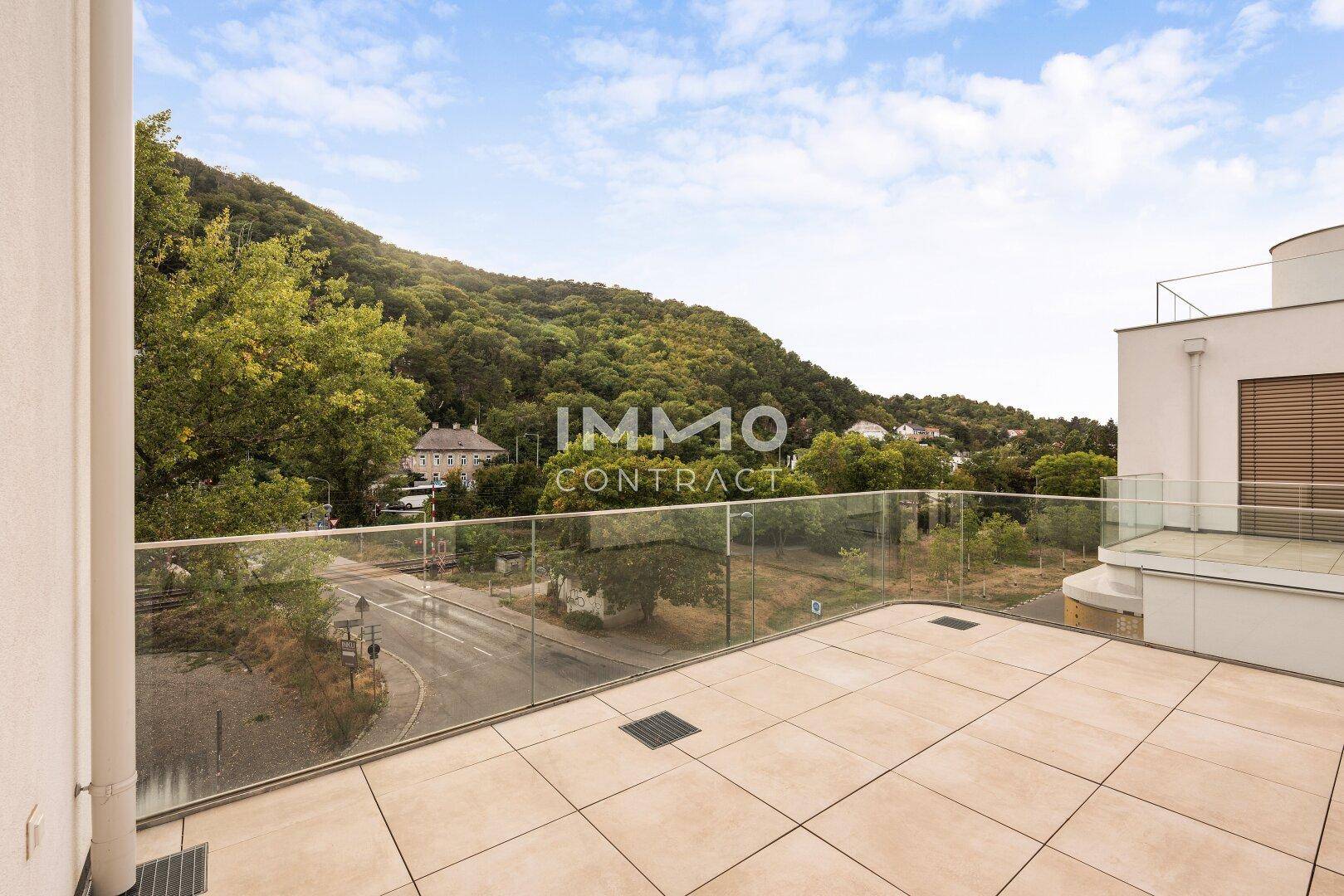 Geräumige Dachterrasse mit Fliesenboden und Glasgeländer, umgeben von grünen Hügeln und Bäumen.