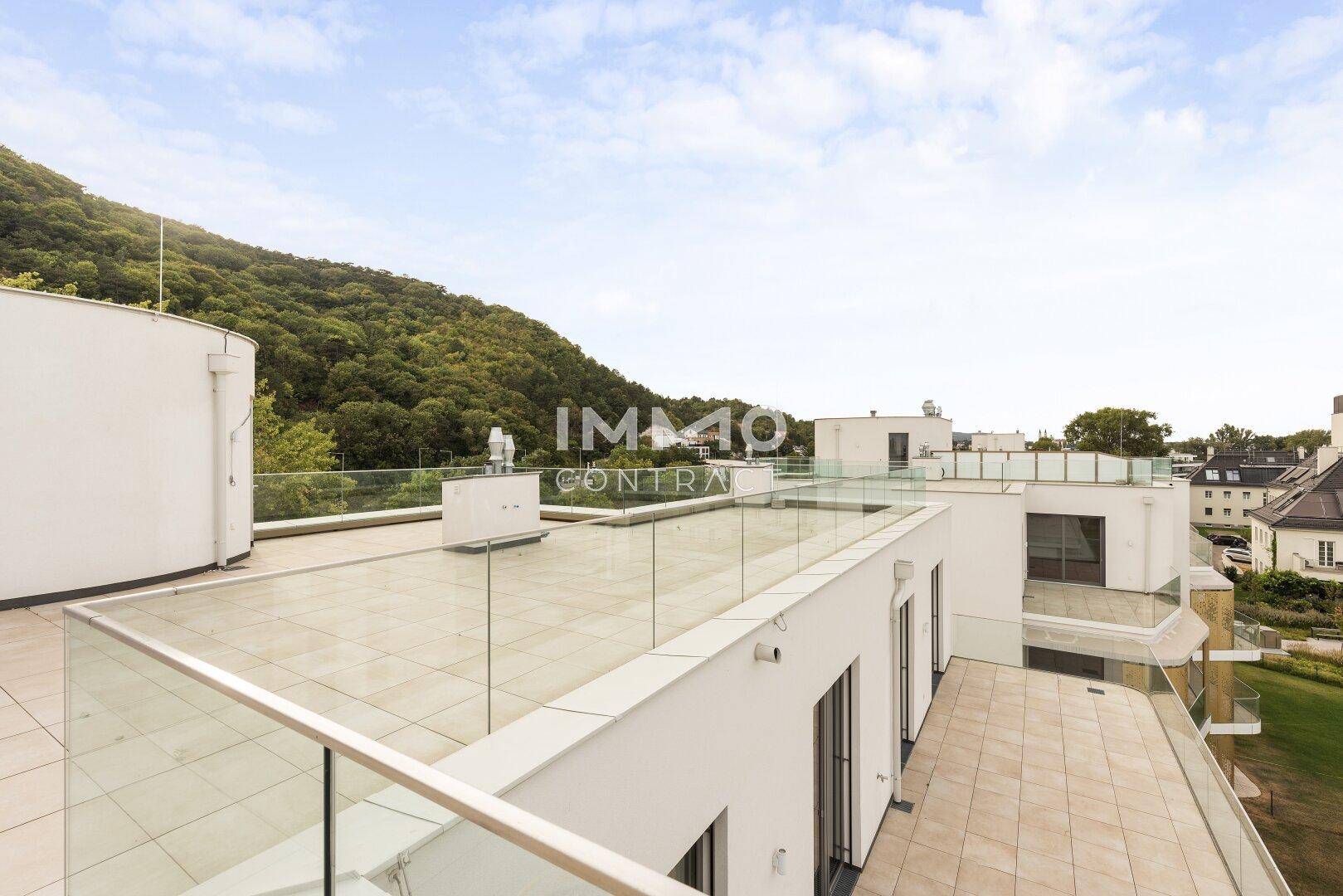 Geräumige Dachterrasse mit Fliesenboden und Glasgeländer, umgeben von grünen Hügeln und Bäumen.