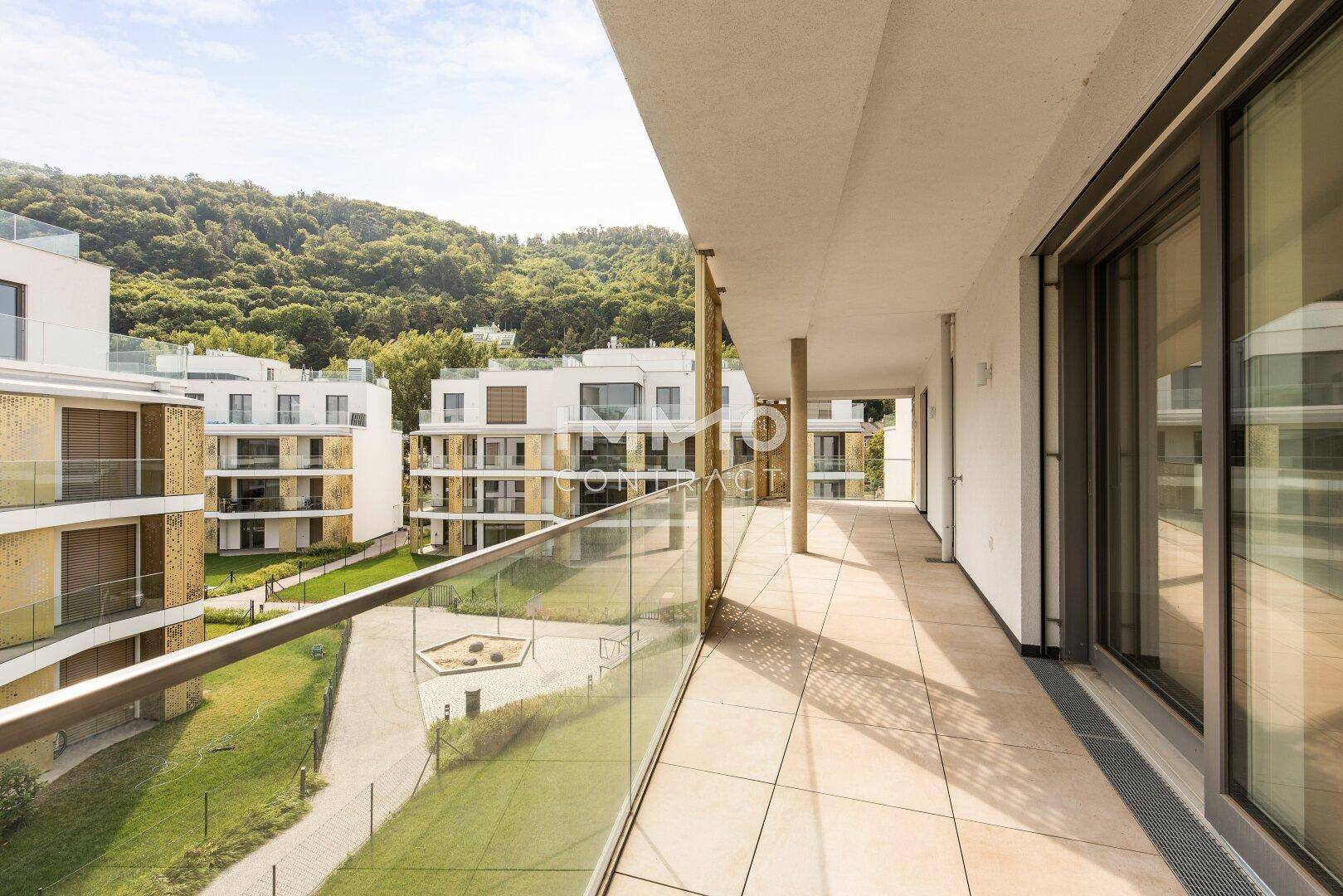 Langer Balkon mit Fliesenboden und Glasgeländer, umgeben von moderner Architektur und grünen Hügeln.