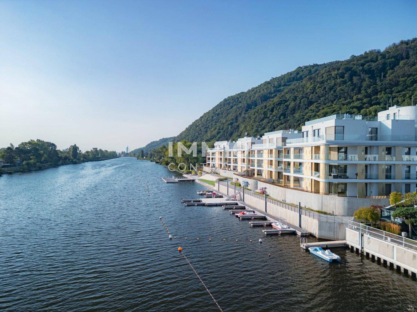Moderne Wohnanlage am Flussufer mit Bootsanlegestellen und grüner Hügellandschaft im Hintergrund.