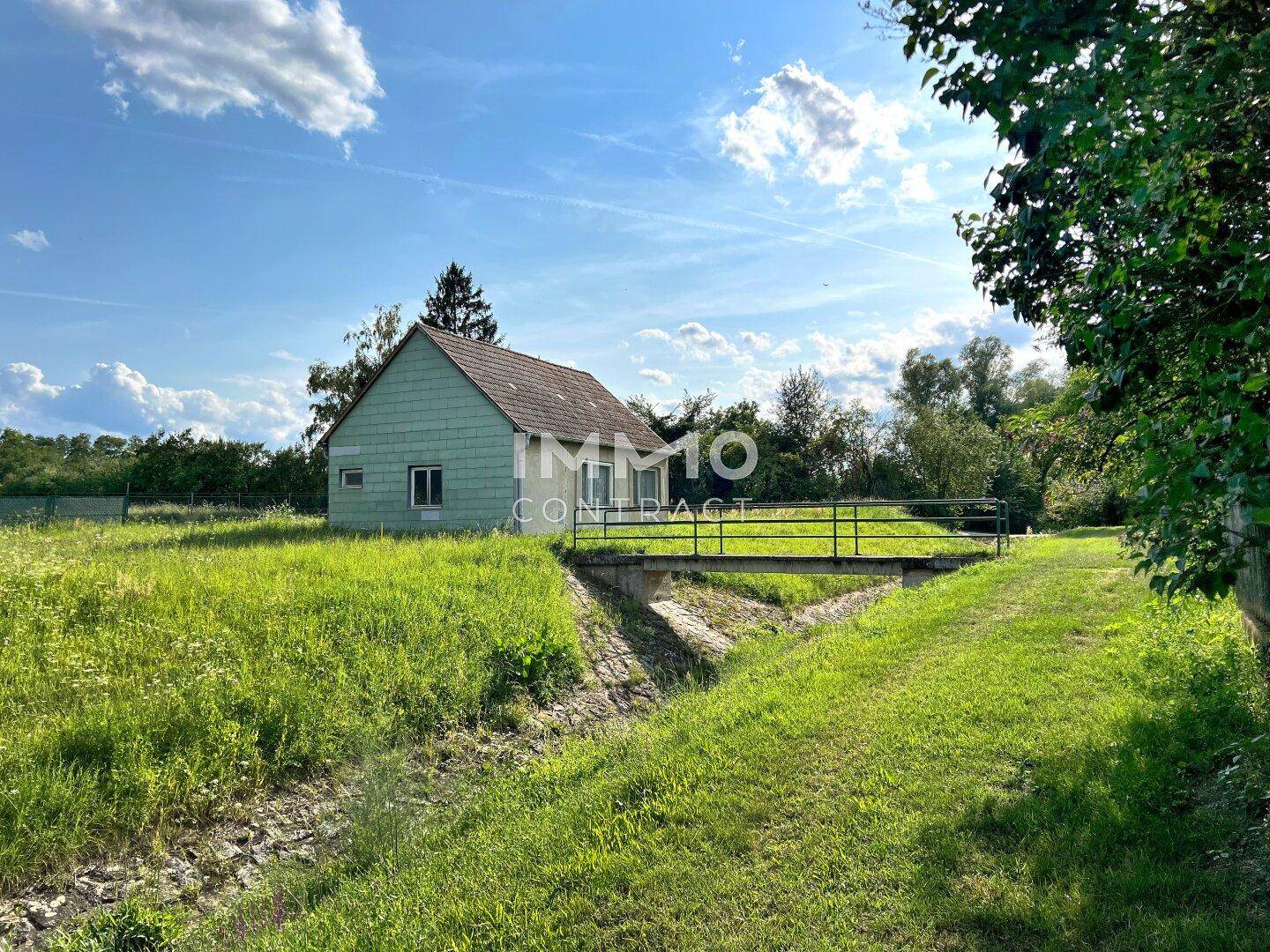 Ländliches Haus mit grüner Fassade, umgeben von Wiesen und einem kleinen Bach mit Brücke.