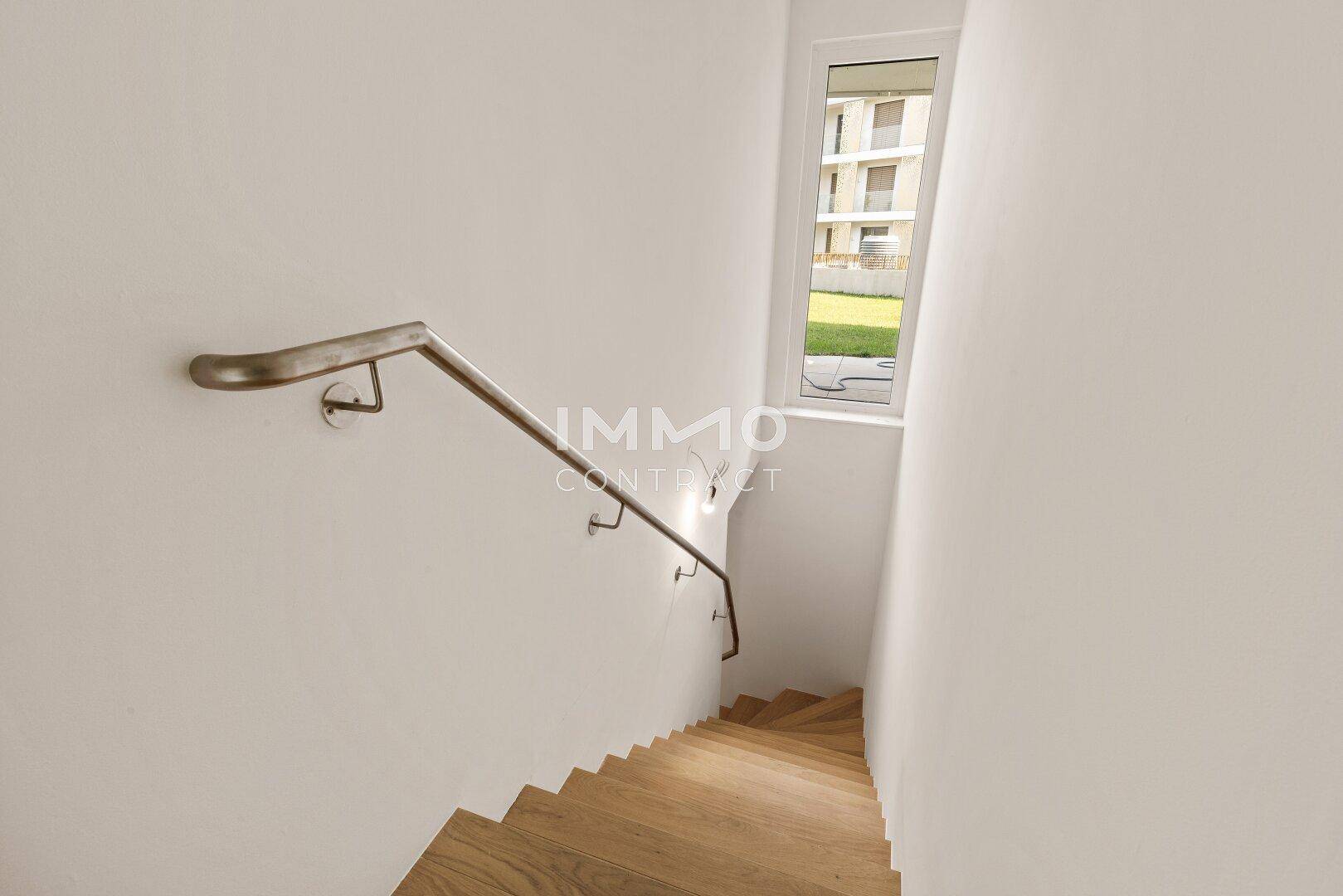 Treppenhaus mit Holzstufen und modernem Handlauf, beleuchtet durch ein schmales Fenster.