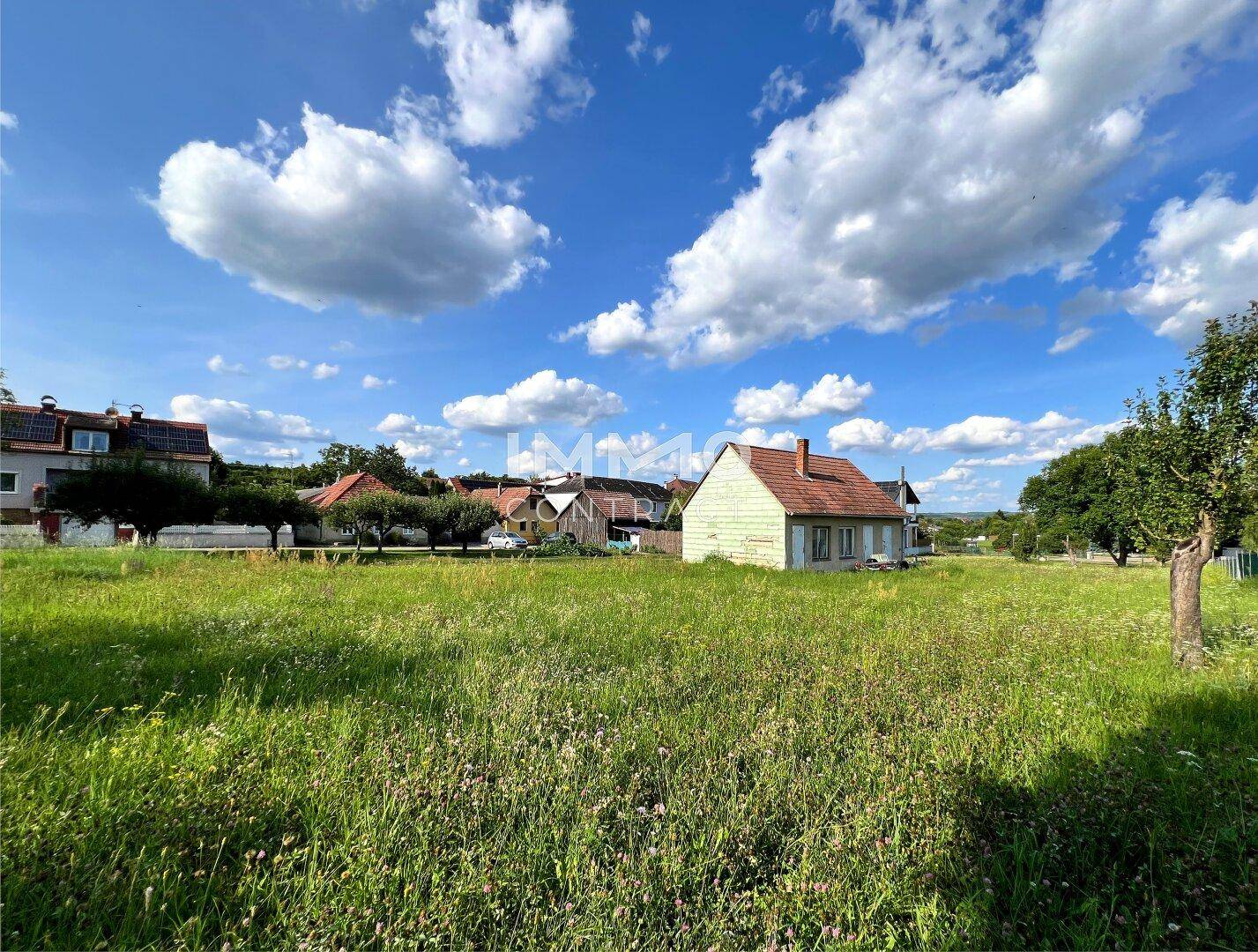 Ländliches Haus in einer weiten Wiese mit weiteren Gebäuden im Hintergrund unter blauem Himmel.