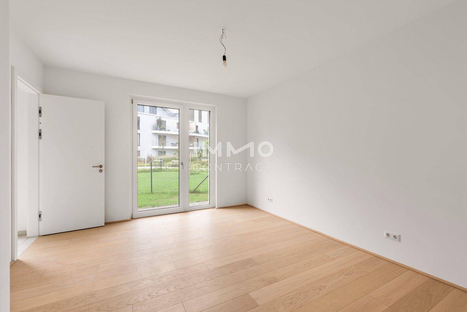 Leeres Schlafzimmer mit hellem Holzboden und Zugang zu einem Außenbereich mit Blick auf andere Gebäude.