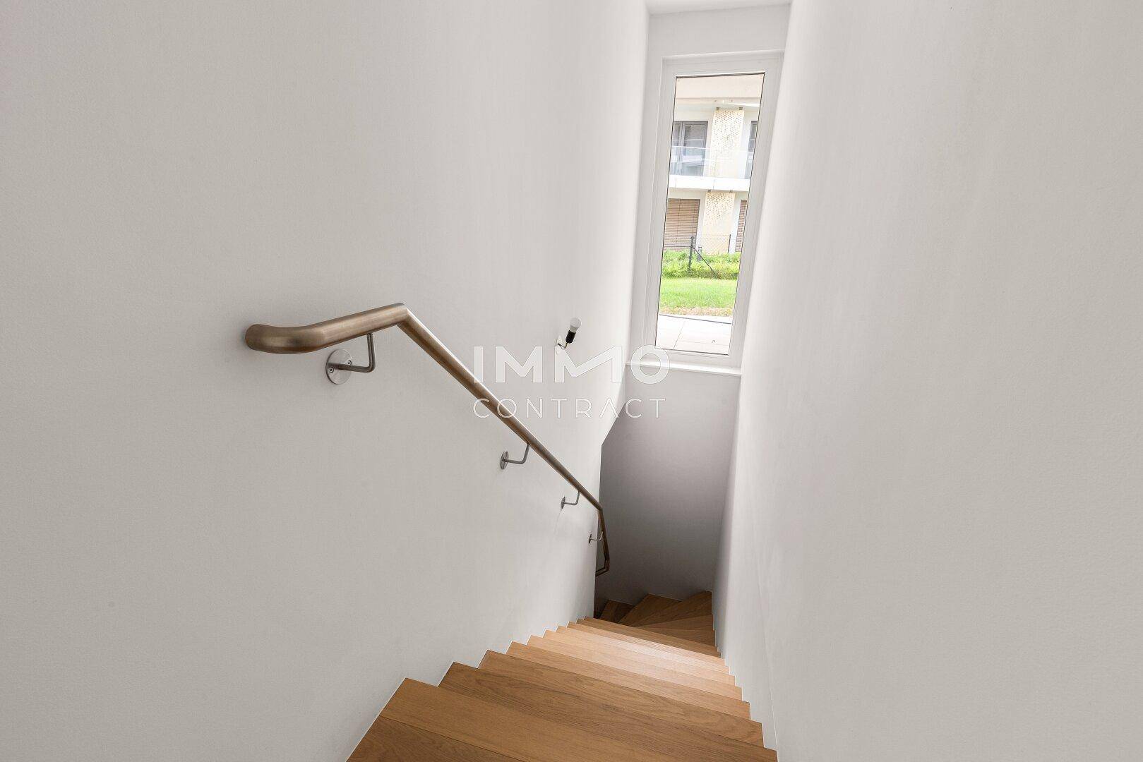 Helles Treppenhaus mit Holzstufen, Metallgeländer und einem Fenster, das Tageslicht hereinlässt.