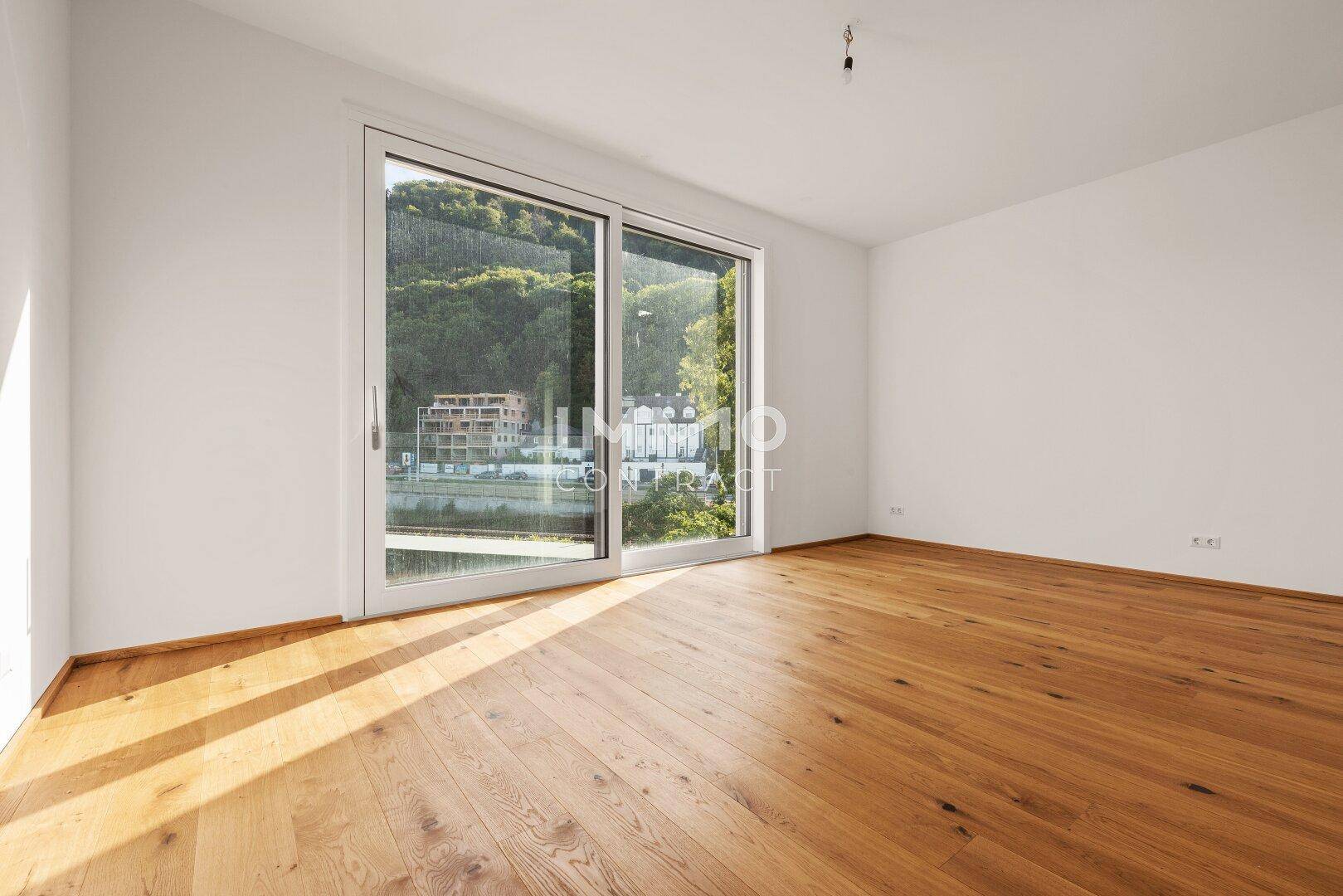 Sonniger Raum mit Holzboden und großem Fenster, das einen weiten Blick auf die Landschaft bietet.