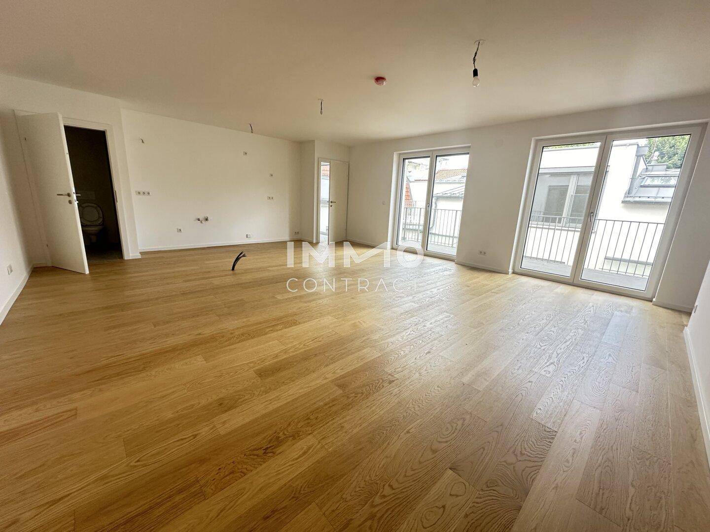 Geräumiger, unmöblierter Wohnbereich mit Holzboden und großen Fenstern zum Balkon, lichtdurchflutet und offen.
