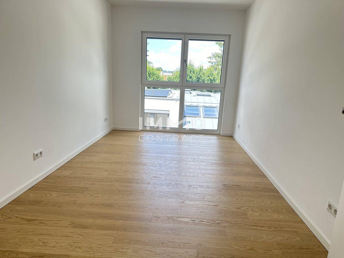 Leeres Schlafzimmer mit Holzboden und großem Fenster mit Blick ins Grüne, bietet viel Tageslicht.