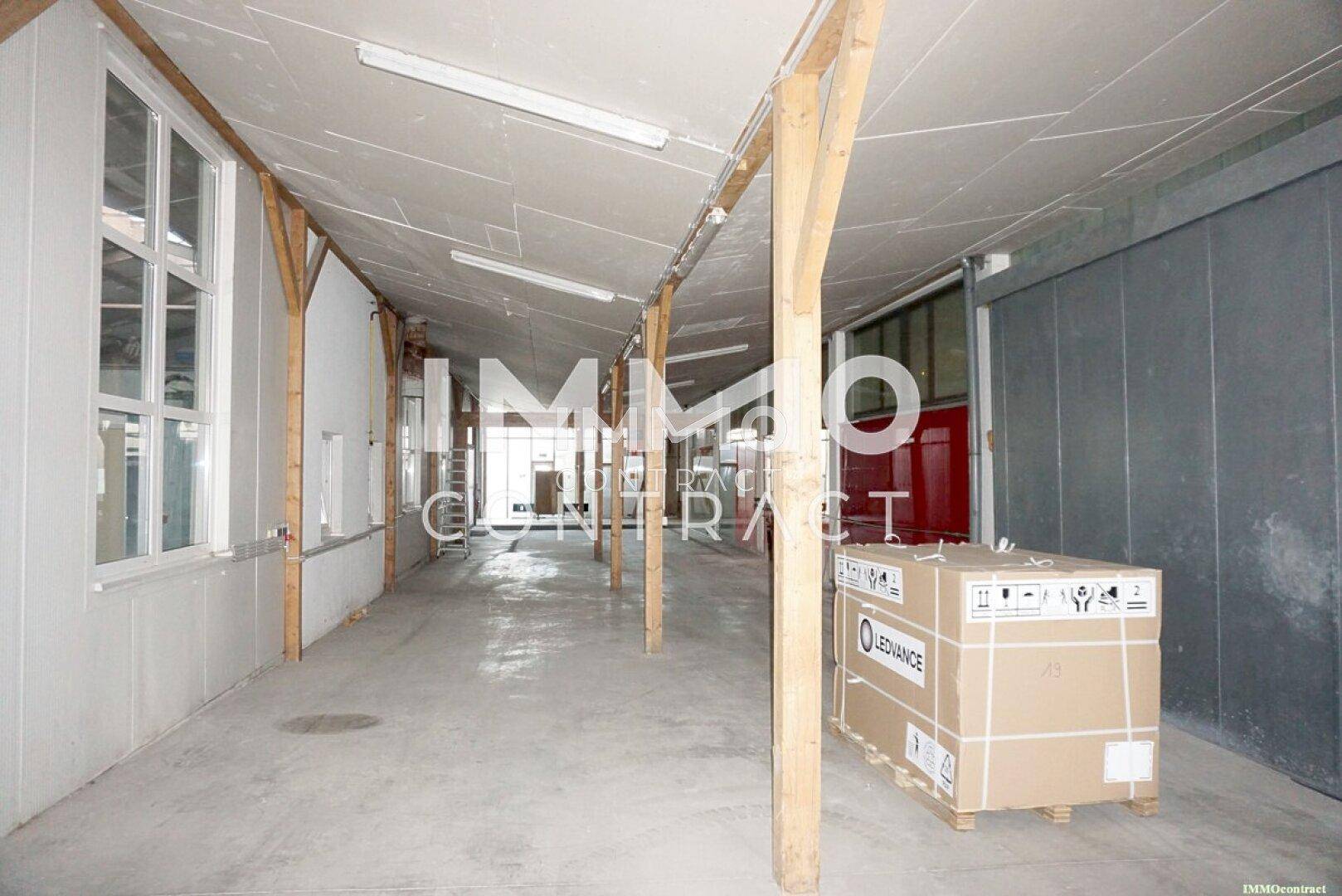 Langer Gang in einer Halle mit Holzbalken, Betonboden und großen Fenstern, gut beleuchtet.