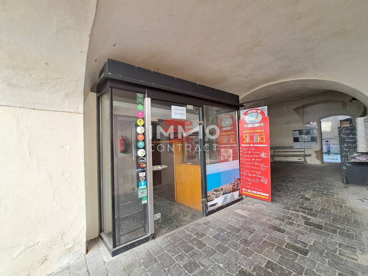 Kleiner Laden unter einem historischen Torbogen mit Glasfront und Werbeschildern, gut sichtbar für Passanten.