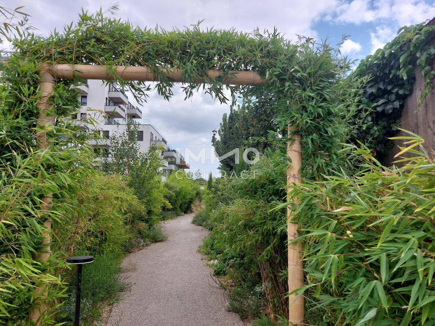 Grüner Gartenweg unter einem bewachsenen Bogen, umgeben von üppiger Vegetation und modernen Gebäuden im Hintergrund.