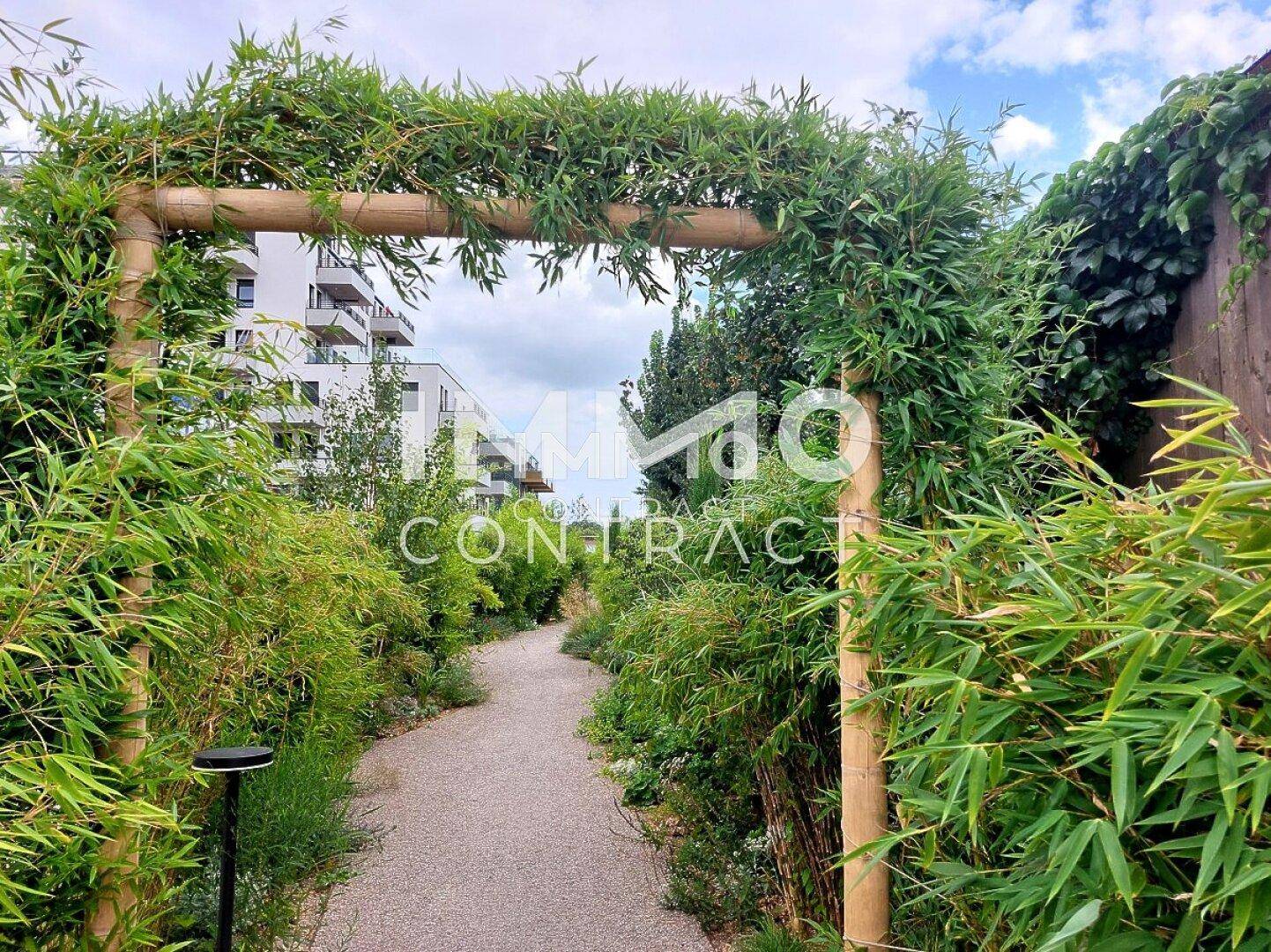Idyllischer Gartenweg mit Bambusbögen und üppiger Vegetation, der zu den Wohngebäuden führt.