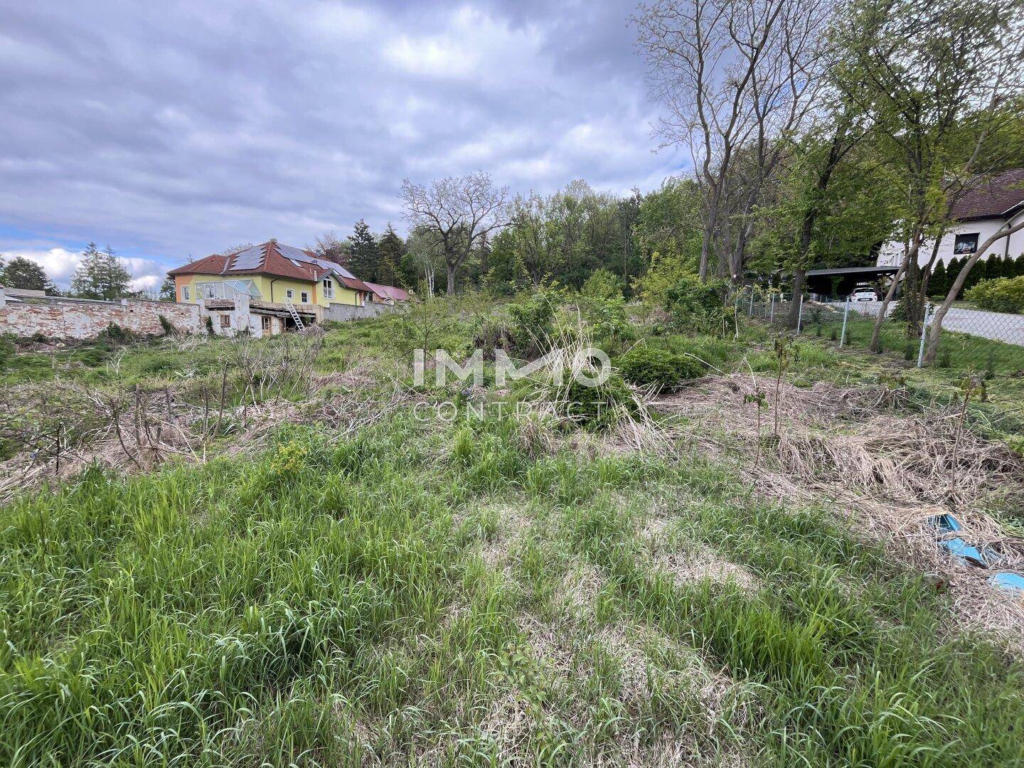 Blick über das ungenutzte Baugrundstück mit einem Wohnhaus und bewaldeten Hügeln im Hintergrund.