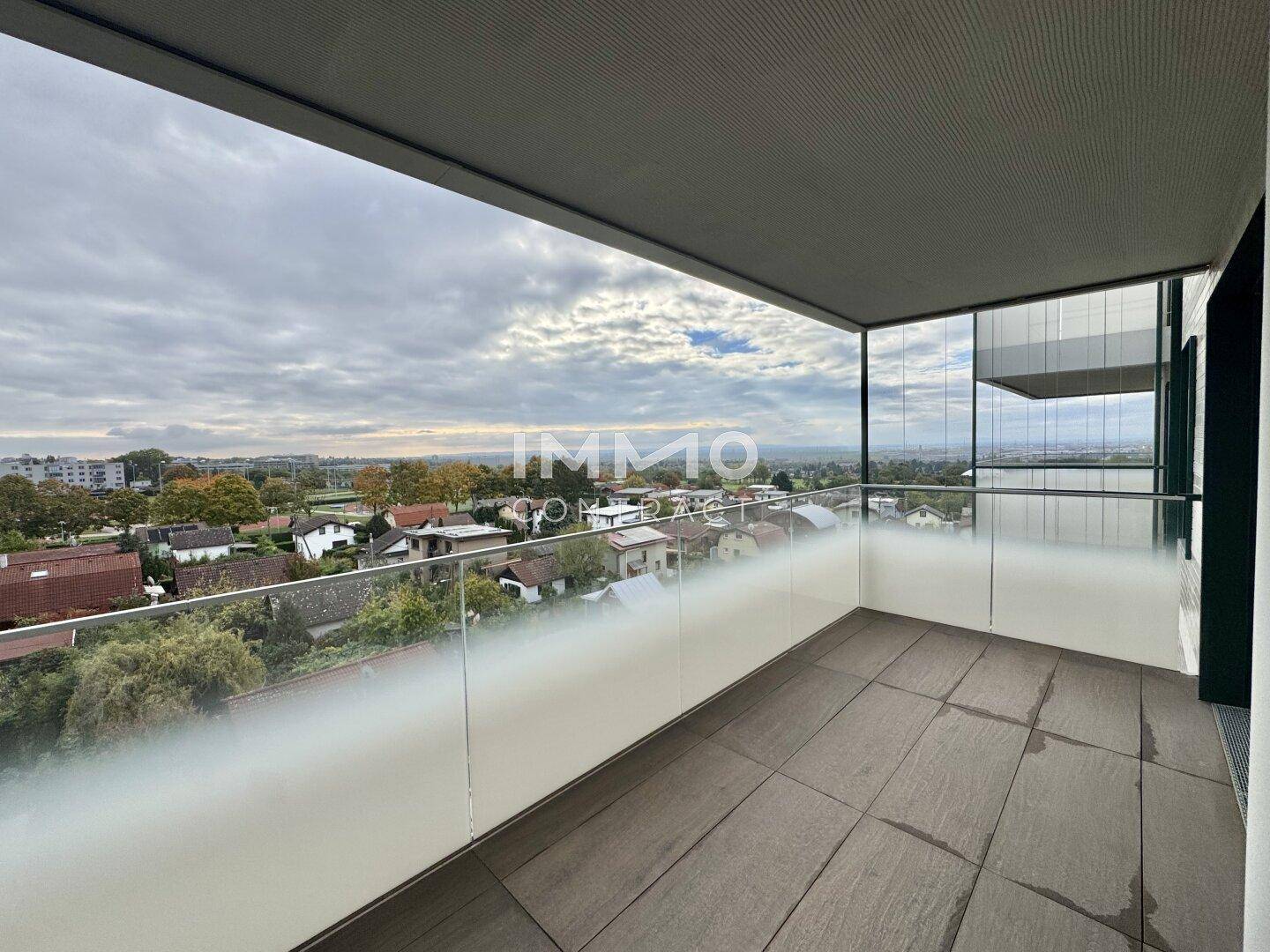 Moderner Balkon mit Glasgeländer und weitem Blick über die Wohngegend unter bewölktem Himmel.