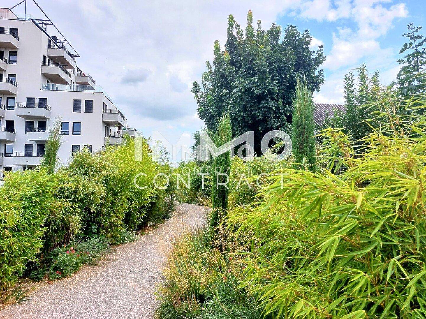 Gepflegter Gartenweg mit vielfältiger Bepflanzung neben einem modernen Wohngebäude mit Balkonen.