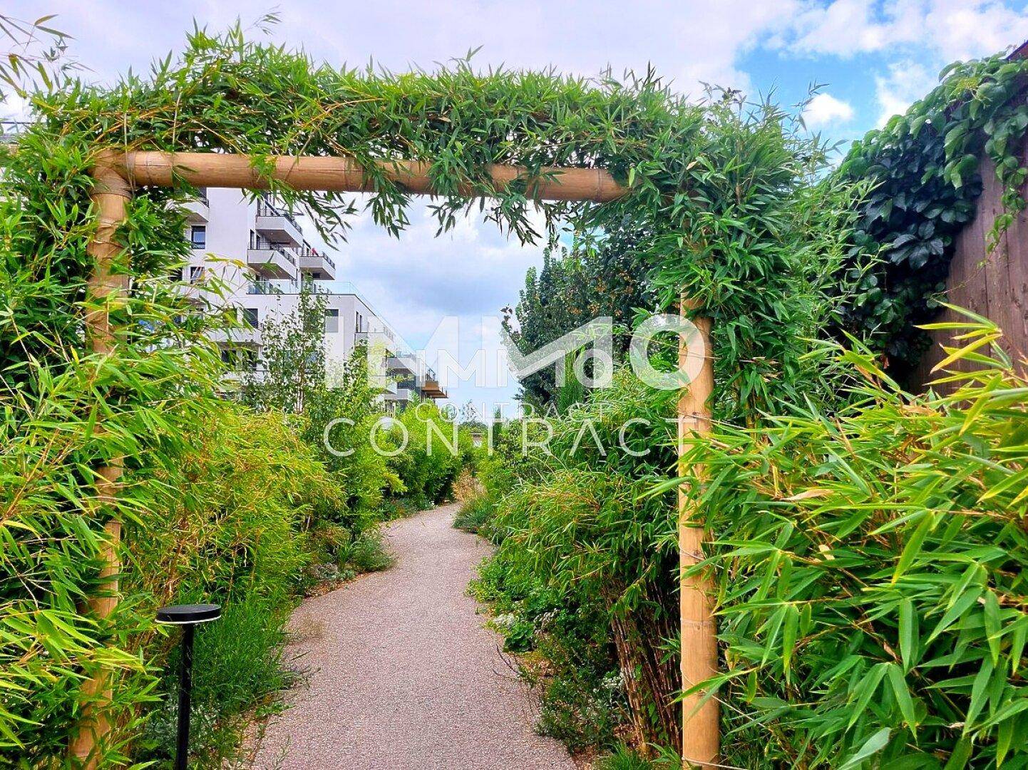 Begrünter Gartenweg mit einem Bambusbogen, der zu einem modernen Wohngebäude im Hintergrund führt.