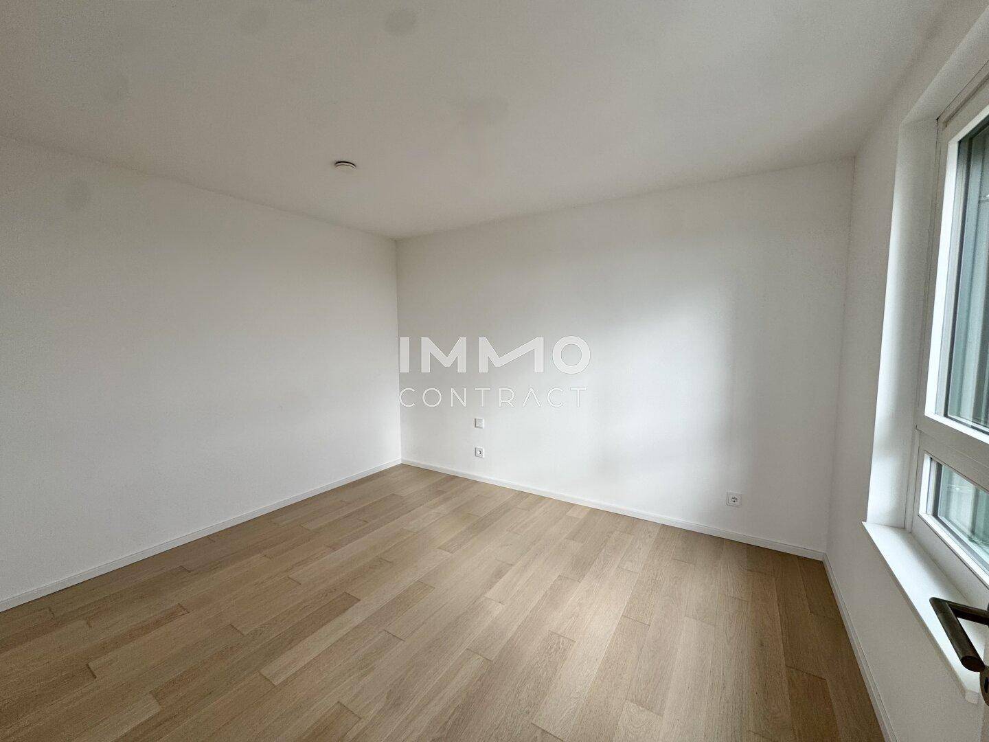 Helles, leeres Zimmer mit Parkettboden und einem Fenster, das viel Tageslicht hereinlässt.