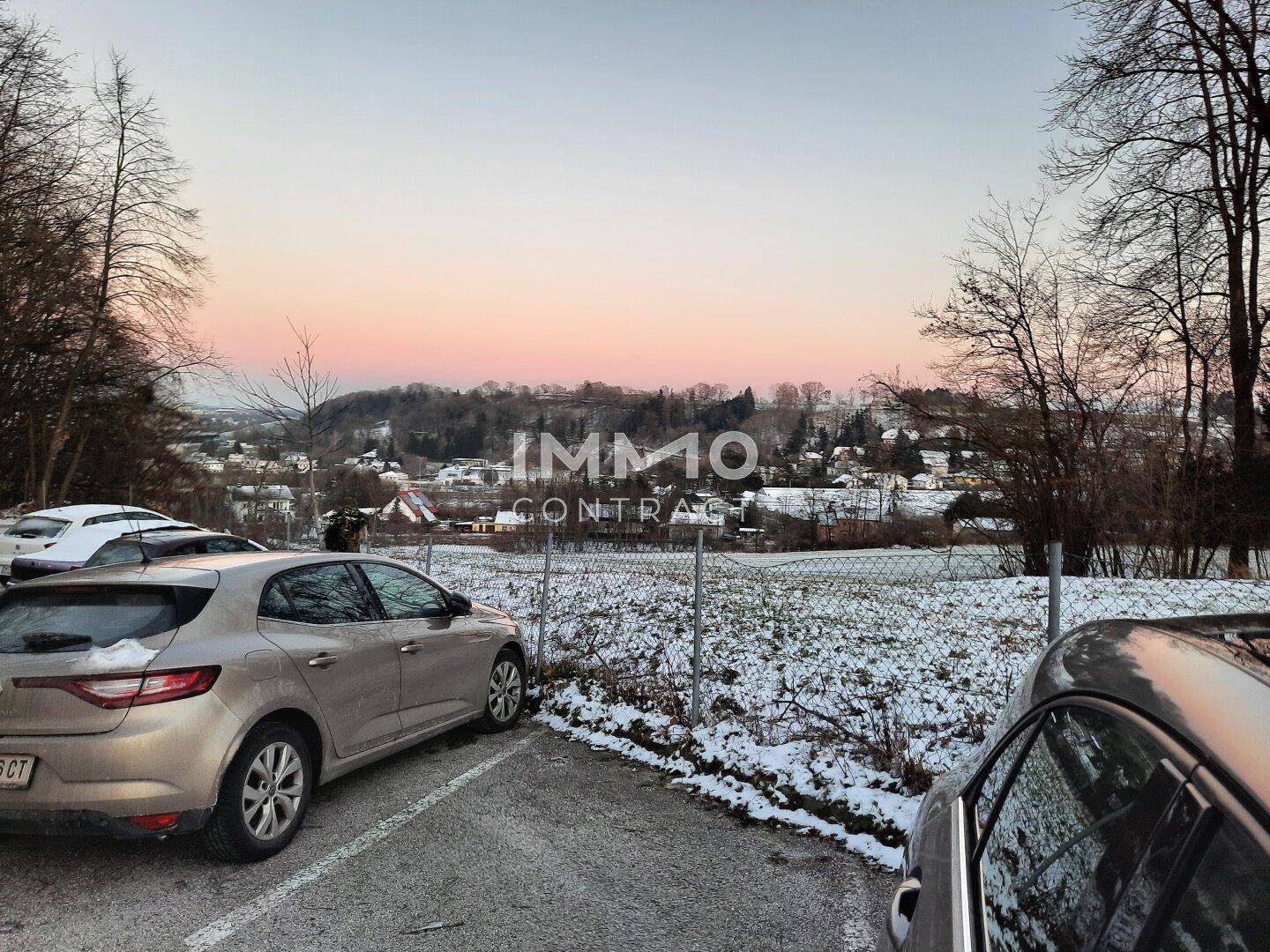 Winterliche Landschaft mit verschneiten Feldern, parkenden Autos und einem weiten Blick auf die Umgebung bei Sonnenuntergang.