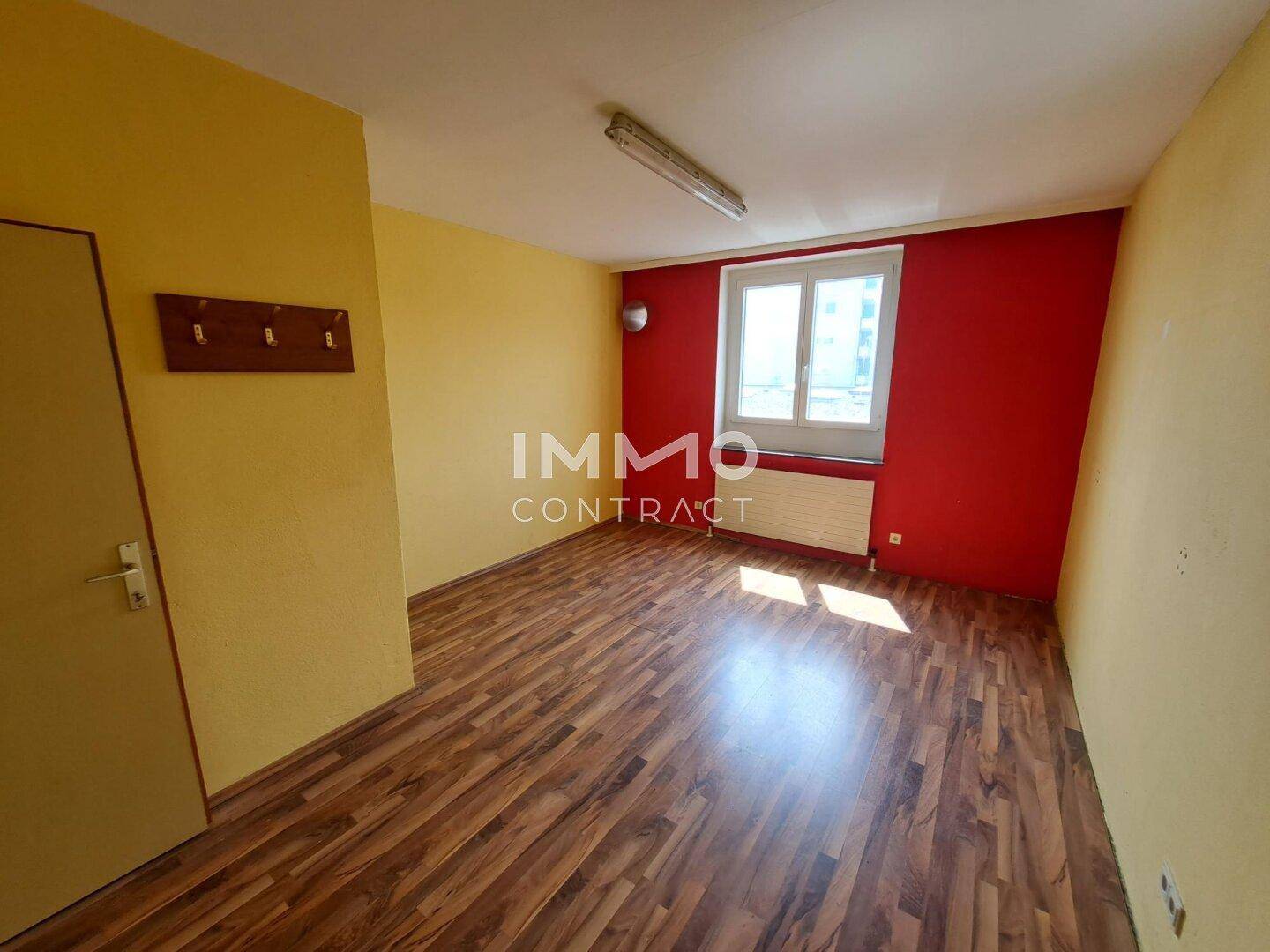 Kleiner Raum mit Holzboden, gelben und roten Wänden sowie einem Fenster mit Heizkörper.