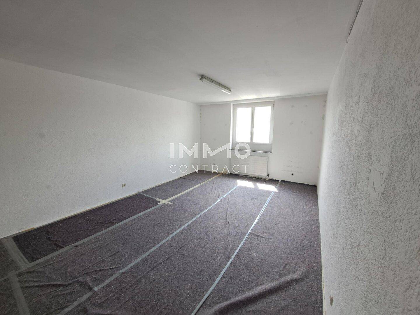 Leerer Raum mit weißen Wänden, einem Fenster und einem dunklen Bodenbelag.