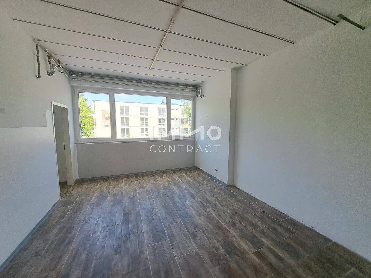Heller Raum mit großen Fenstern, Holzboden und Blick auf andere Gebäude in der Umgebung.