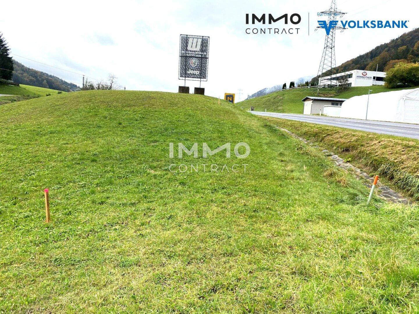 Grüner Hügel mit Werbetafel und Strommast, angrenzend an eine Straße mit Markierungen im Gras.