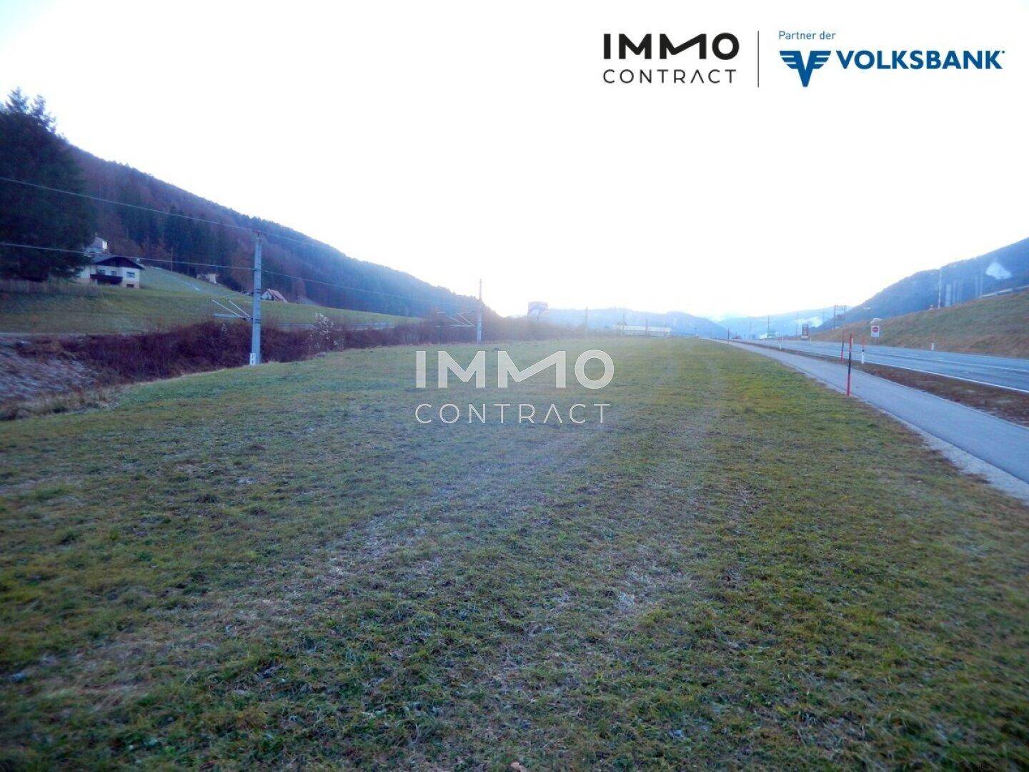 Weitläufiges Grundstück mit spärlicher Vegetation neben einer Straße, umgeben von bewaldeten Bergen.