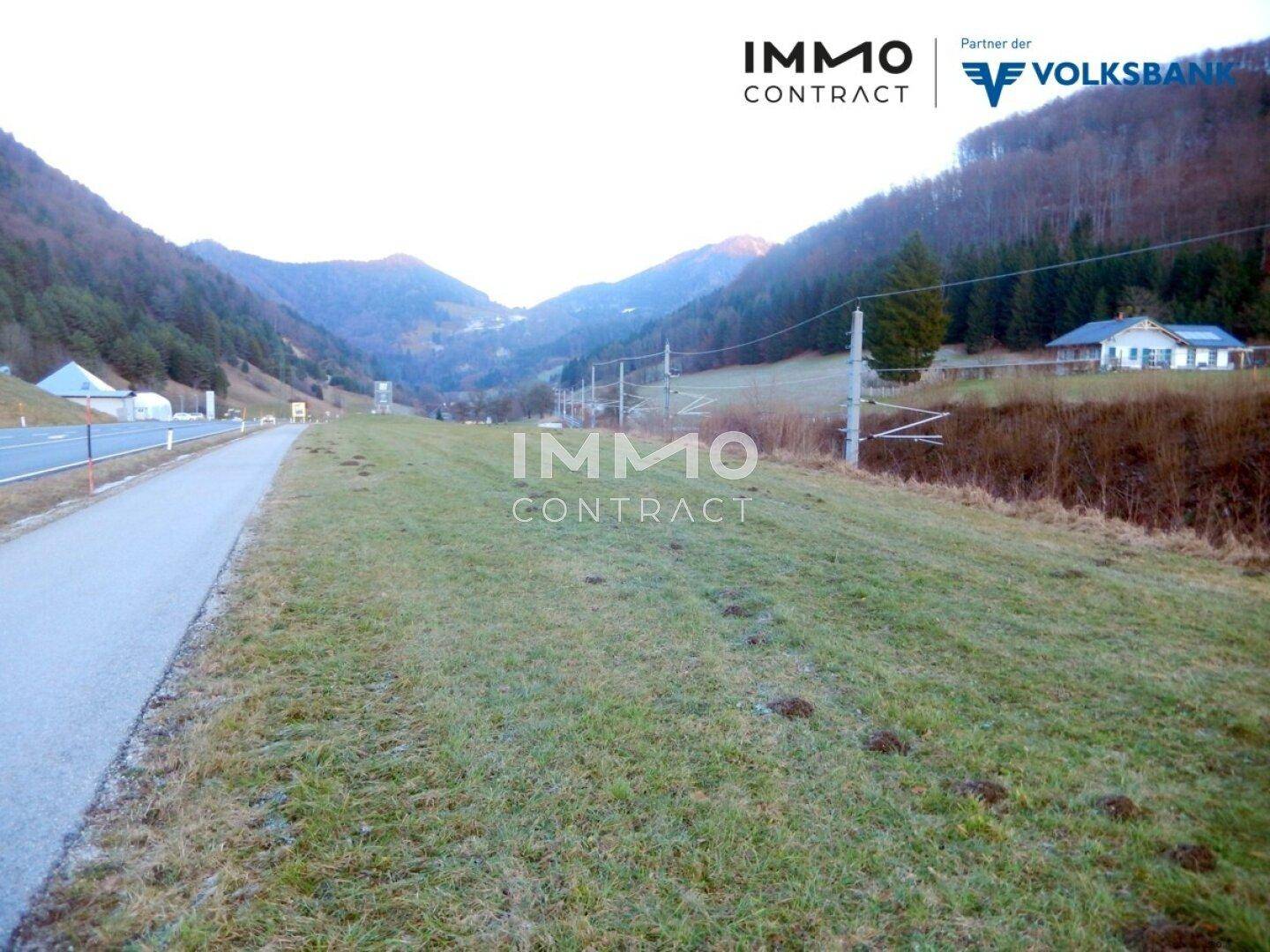 Grünes Grundstück entlang einer Straße mit Häusern und bewaldeten Hügeln in der Ferne.