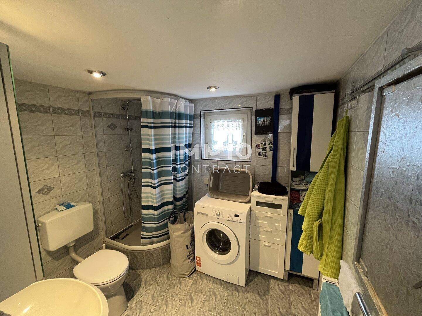 Gefliestes Badezimmer mit Dusche, Toilette, Waschmaschine und einem kleinen Fenster.