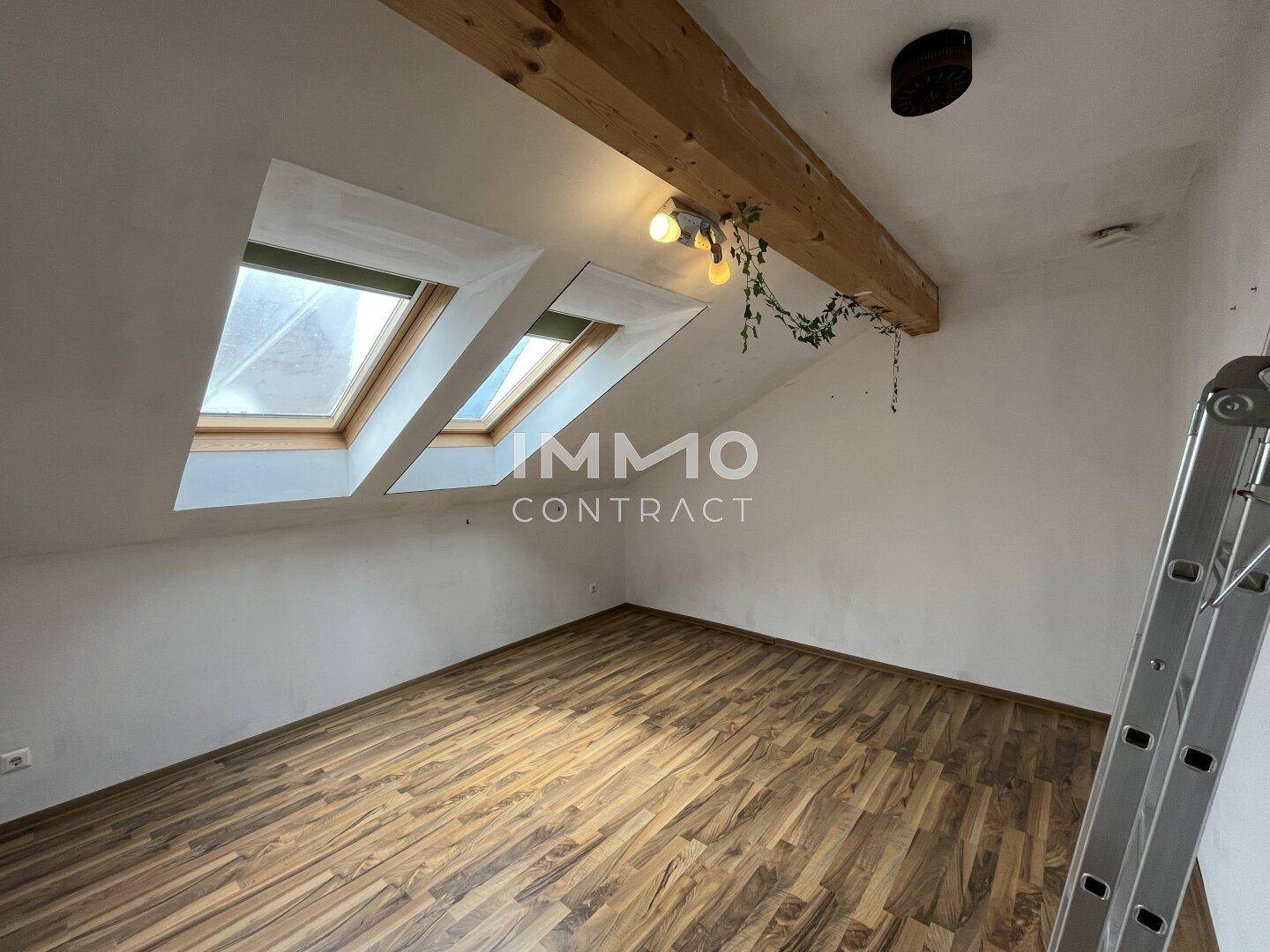Helles Zimmer im Dachgeschoss mit Holzbalken, zwei Dachfenstern und Laminatboden.