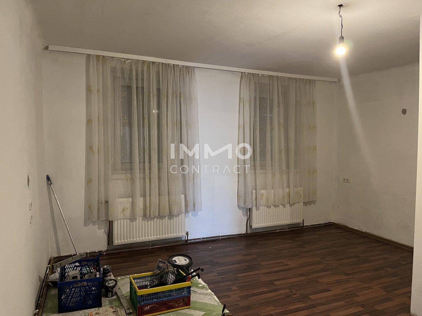 Leeres Zimmer mit Laminatboden, zwei Fenstern und Heizkörpern unter den Fenstern.