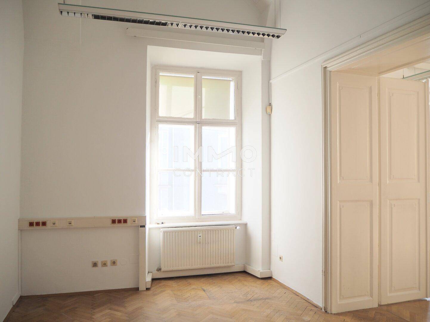 Leerer Büroraum mit Fischgrätparkett, großem Fenster und Heizkörper unter der Fensterbank, ideal für Arbeitsplätze.
