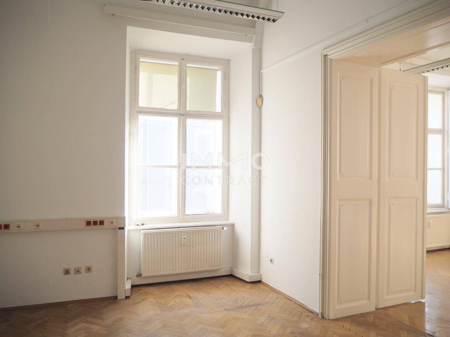 Heller Büroraum mit Fischgrätparkett, hohen Fenstern und Heizkörper unter der Fensterbank, bietet viel Tageslicht.