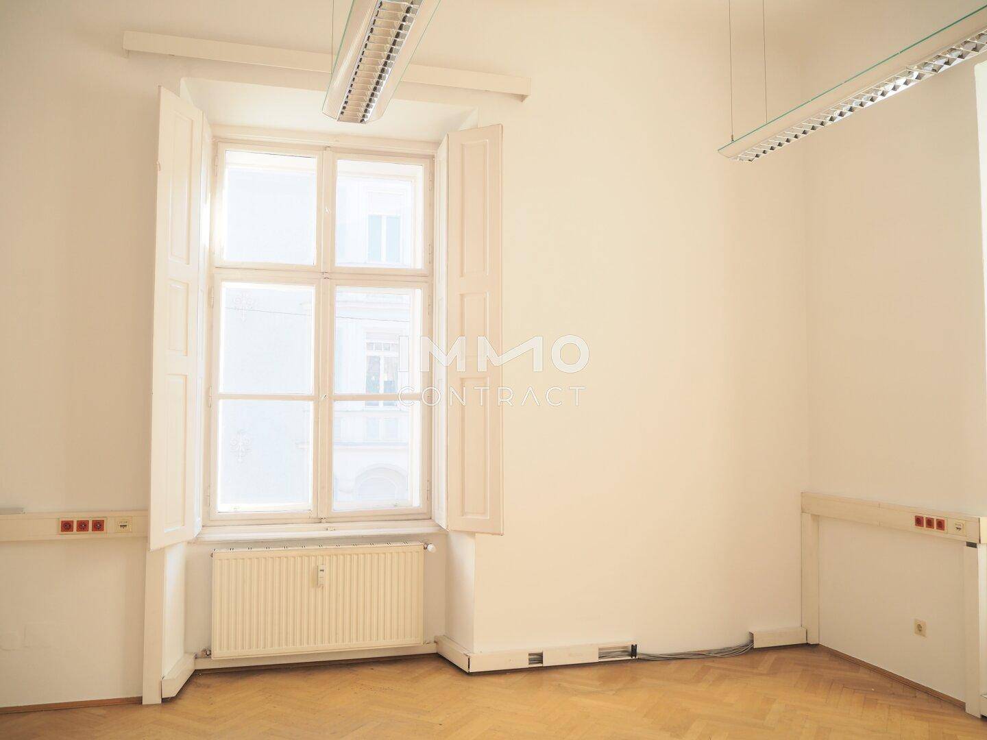 Leerer Büroraum mit Fischgrätparkett, hohen Fenstern mit Fensterläden und Deckenbeleuchtung, sehr hell.
