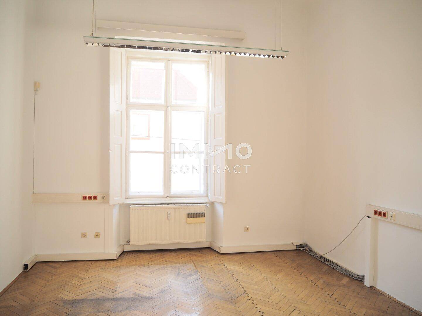 Heller Büroraum mit Fischgrätparkett, hohen Fenstern und Deckenbeleuchtung, schafft eine angenehme Arbeitsatmosphäre.